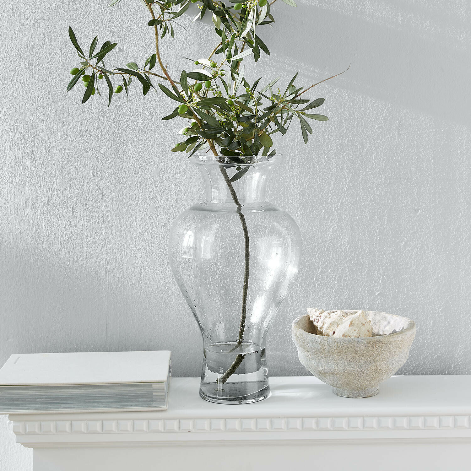 Transparante glazen vaas met groene takken op witte plank met boek en schaal met schelpen