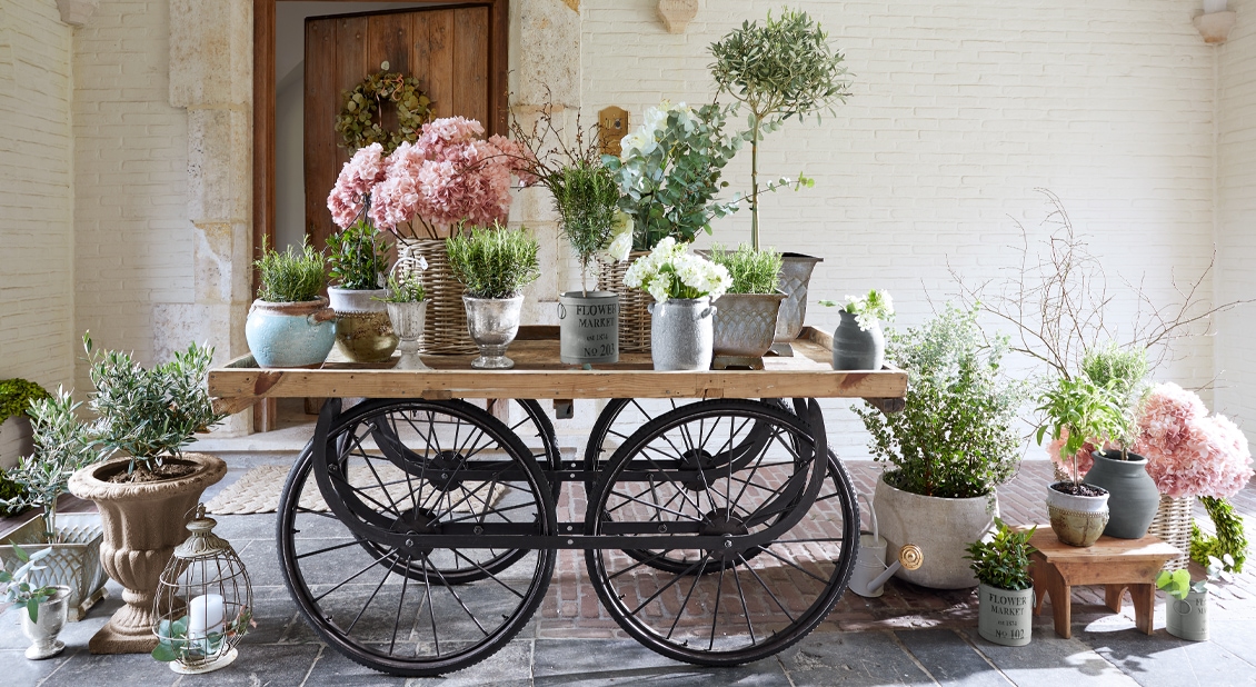 Houten kar met zwarte wielen versierd met potten met roze en witte bloemen en groene planten