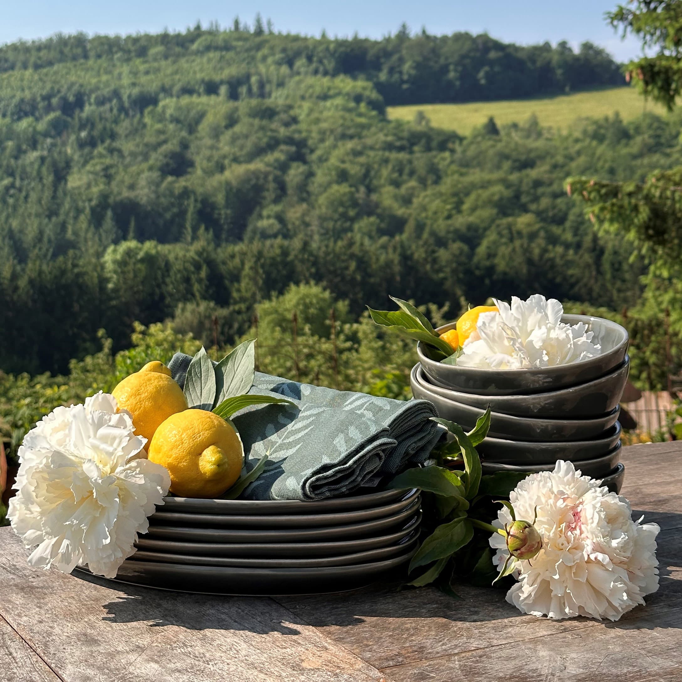 Set grijze keramische borden en kommen met witte bloemen en citroenen op houten tafel met uitzicht op groen bos
