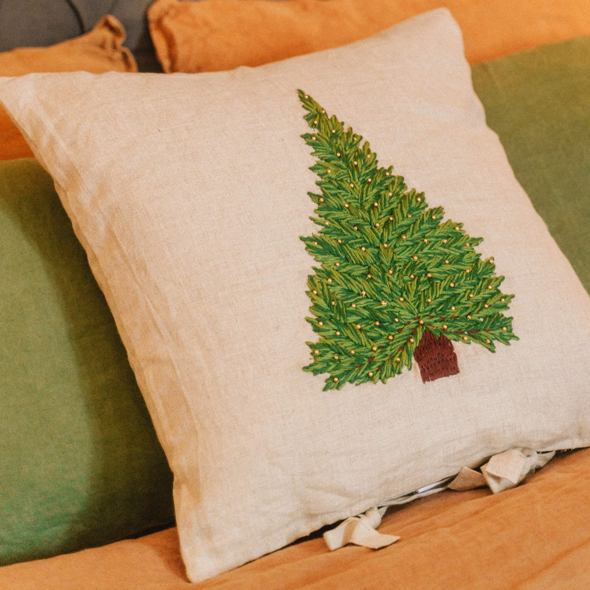 Beige linnen kussen met geborduurde groene kerstboom op oranje en groene beddengoed voor sfeervolle kamer