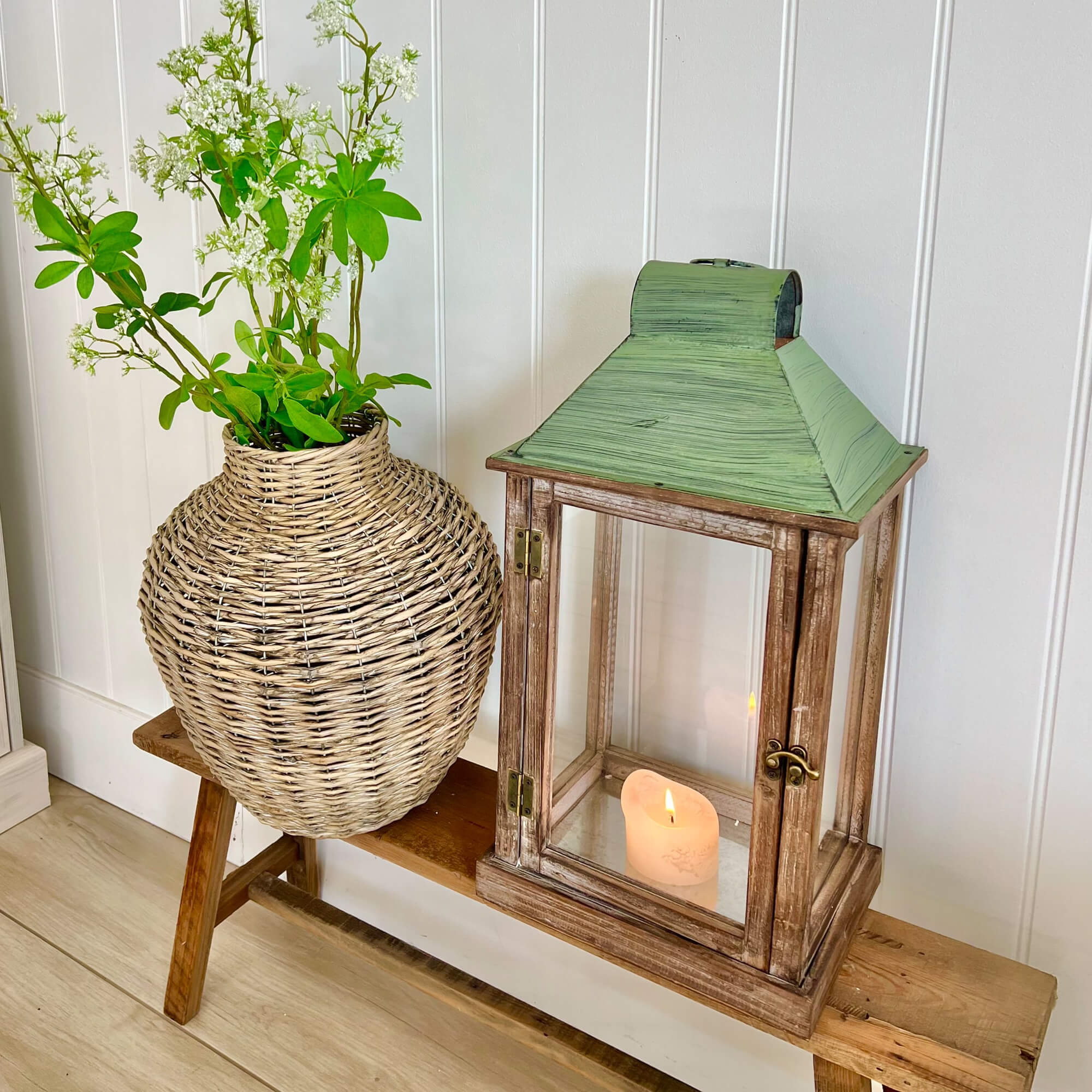 Rustieke houten lantaarn met groen metalen dak en rieten vaas met witte bloemen op houten bank
