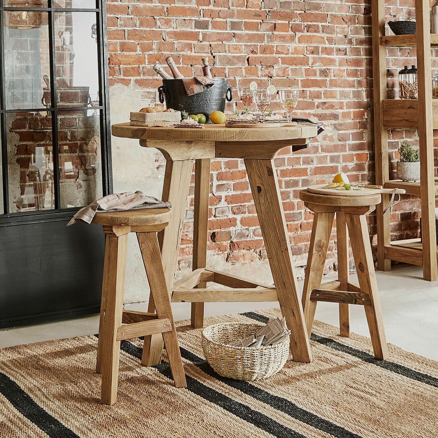 Zitgroep Stonington Hoge houten tafel met twee krukken in natuurlijke kleur op jute vloerkleed in industriële ruimte