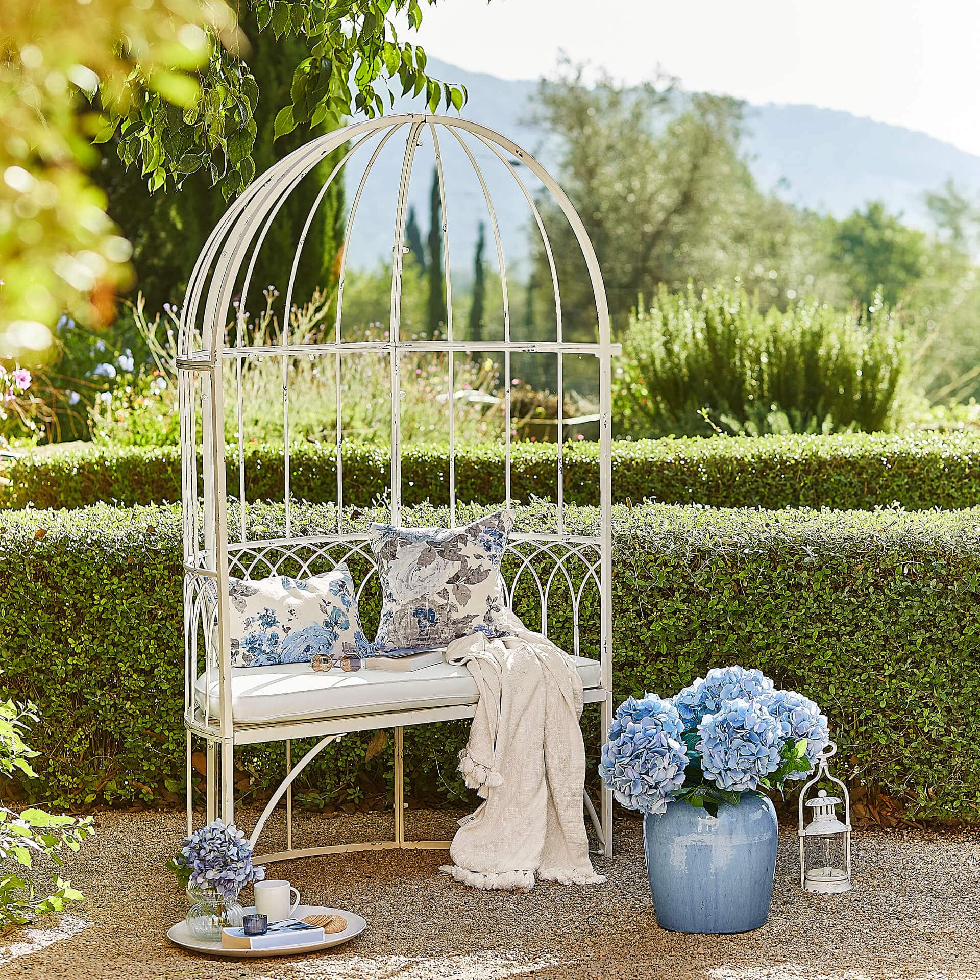 Witte metalen tuinbank met kussens en blauwe bloemen in pot tegen groene achtergrond