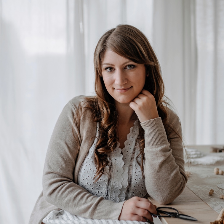 Portret van een vrouw in een beige trui en witte kanten blouse zittend aan een tafel in een lichte kamer