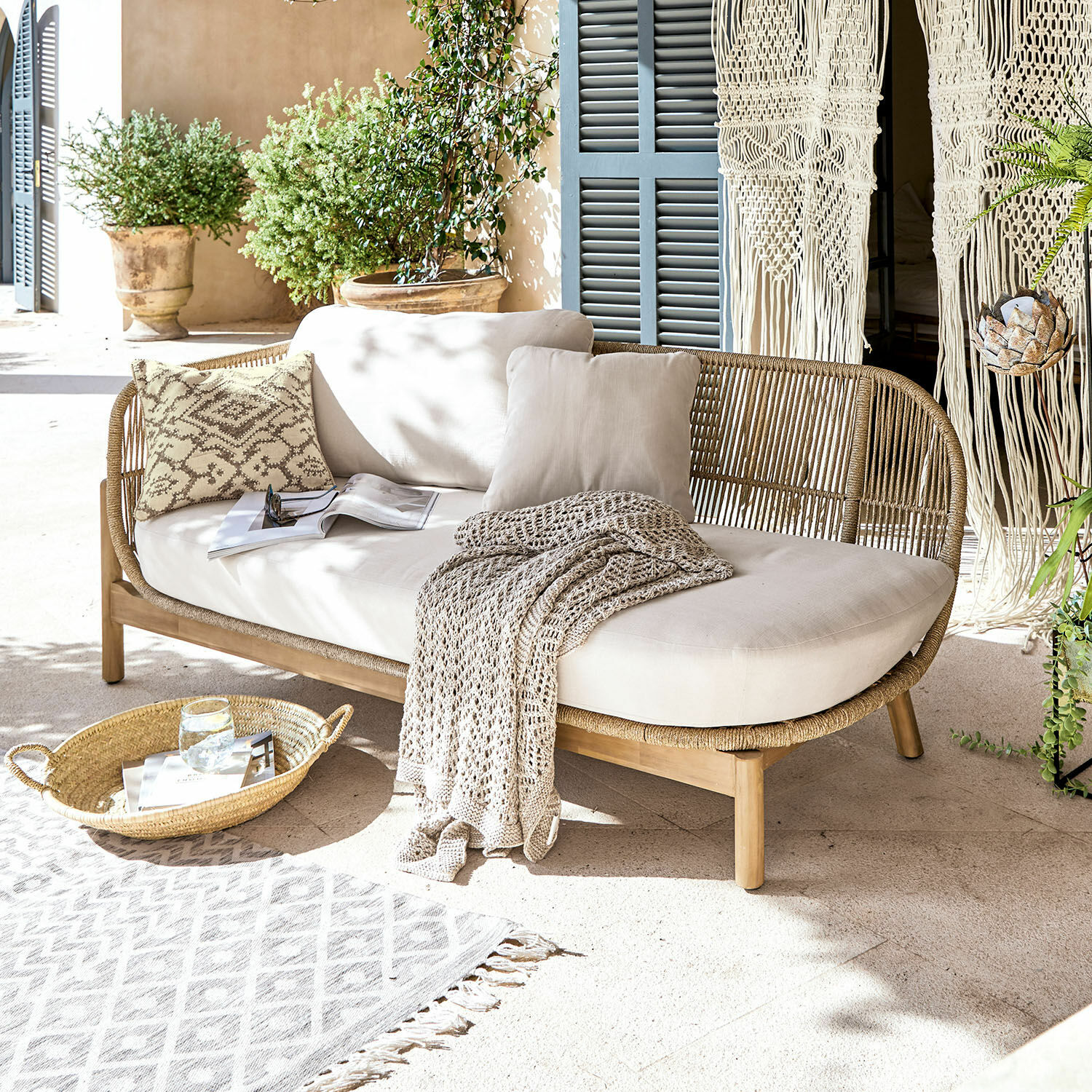 Chaise longue FSC®-100% Belvidere Rotan- en houten bank met crèmekussens en deken op lichte terras met planten