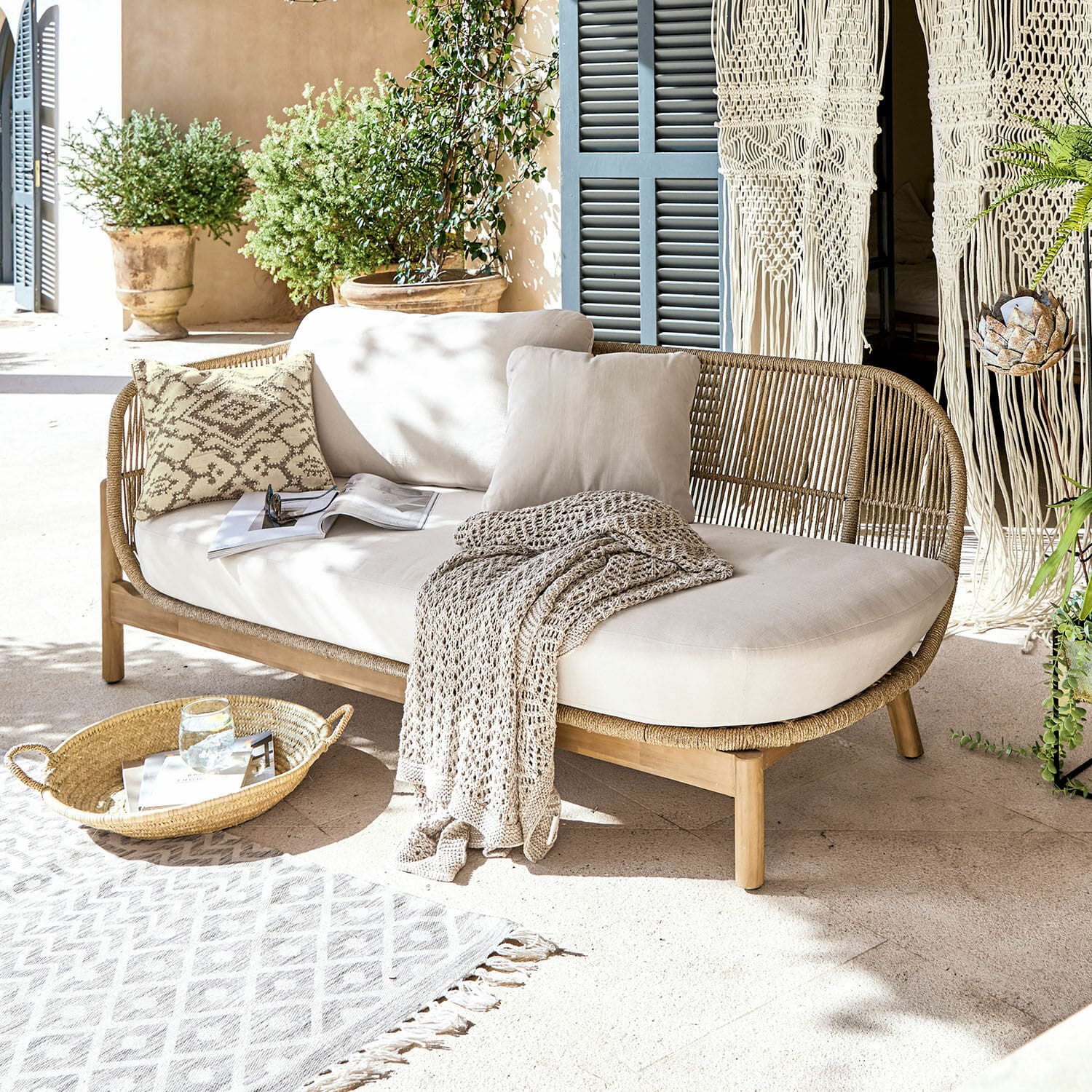 Chaise longue FSC®-100% Belvidere Rotan- en houten bank met crèmekussens en deken op lichte terras met planten