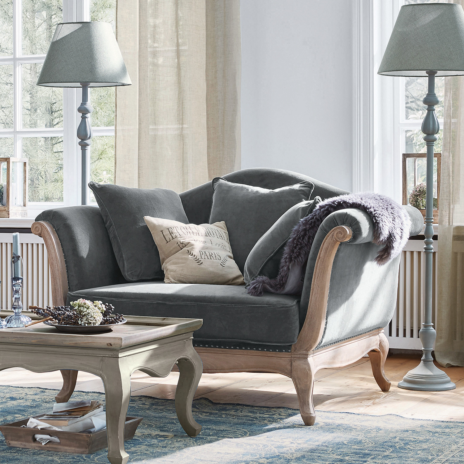 Fauteuil Sumerset Grijze fluwelen bank met houten frame en kussens in lichte woonkamer met staande lampen
