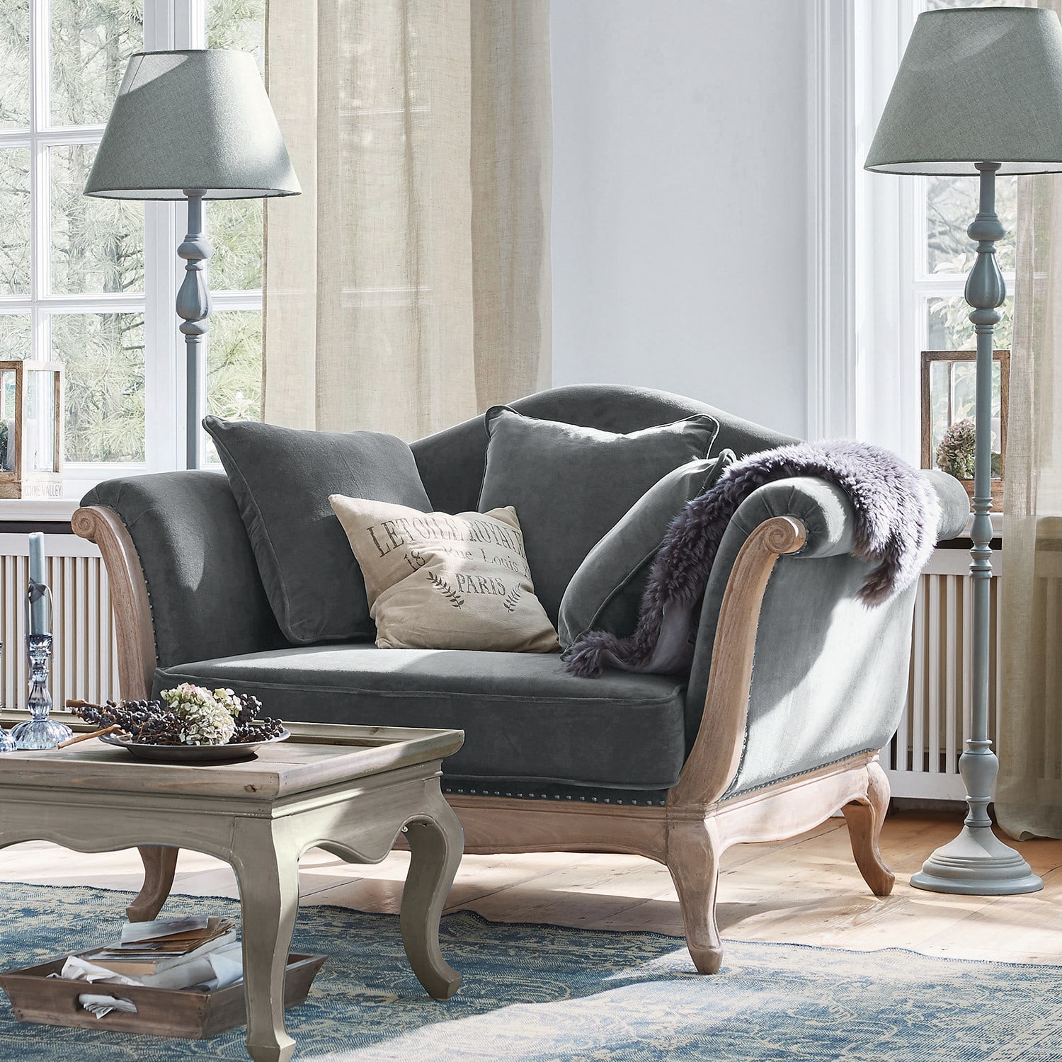 Fauteuil Sumerset Grijze fluwelen bank met houten frame en kussens in lichte woonkamer met staande lampen