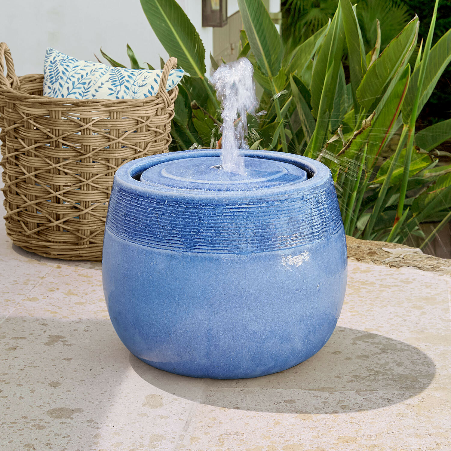 Blauwe keramische tuinfontein met spuitend water, omringd door groene planten en een rieten mand