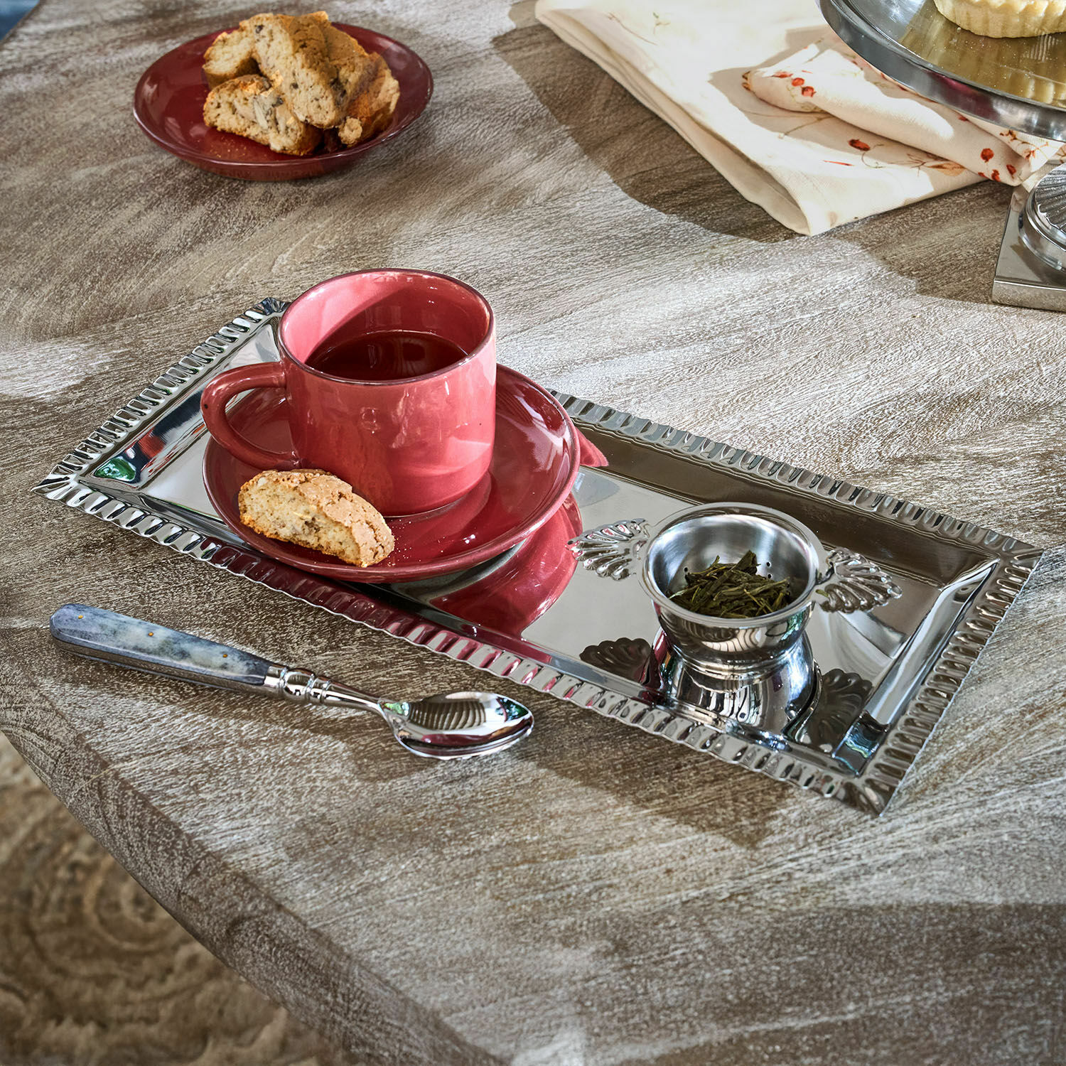 Rechthoekig zilverkleurig metalen dienblad met rode kop, thee en koekje op houten tafel