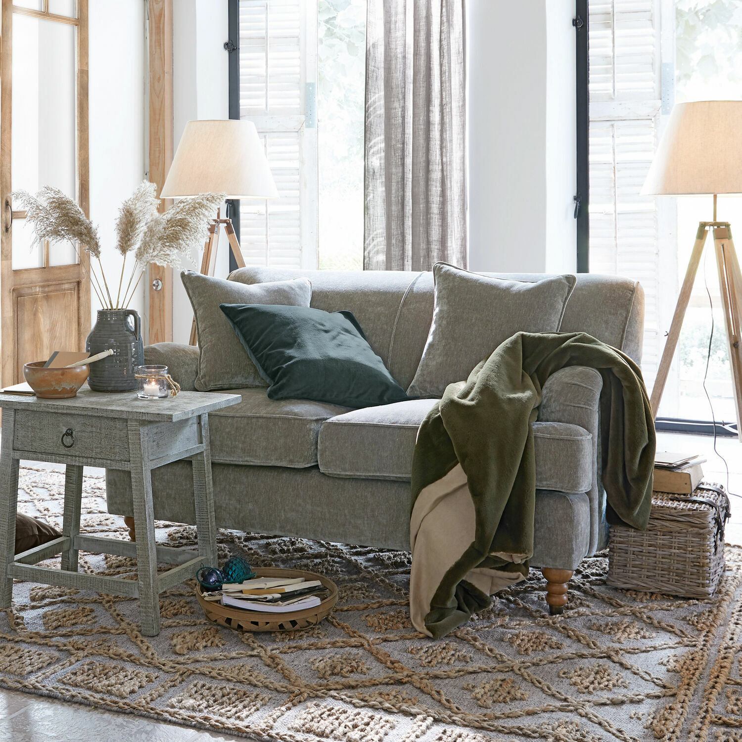 Grijze bank met zachte kussens en groene deken in lichte woonkamer met houten lampen en vloerkleed
