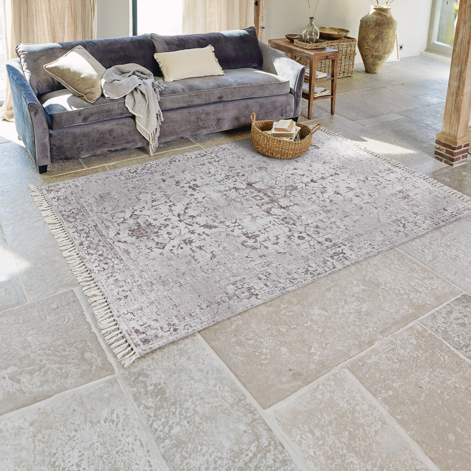 Grijs vloerkleed met subtiel patroon en franjes op stenen vloer in lichte woonkamer met grijze bank