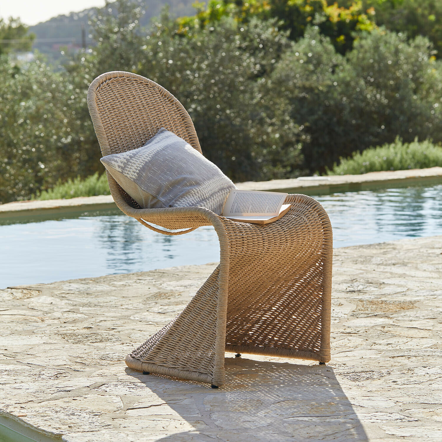 Tuinstoel van natuurlijk vlechtwerk met kussen en boek bij het zwembad