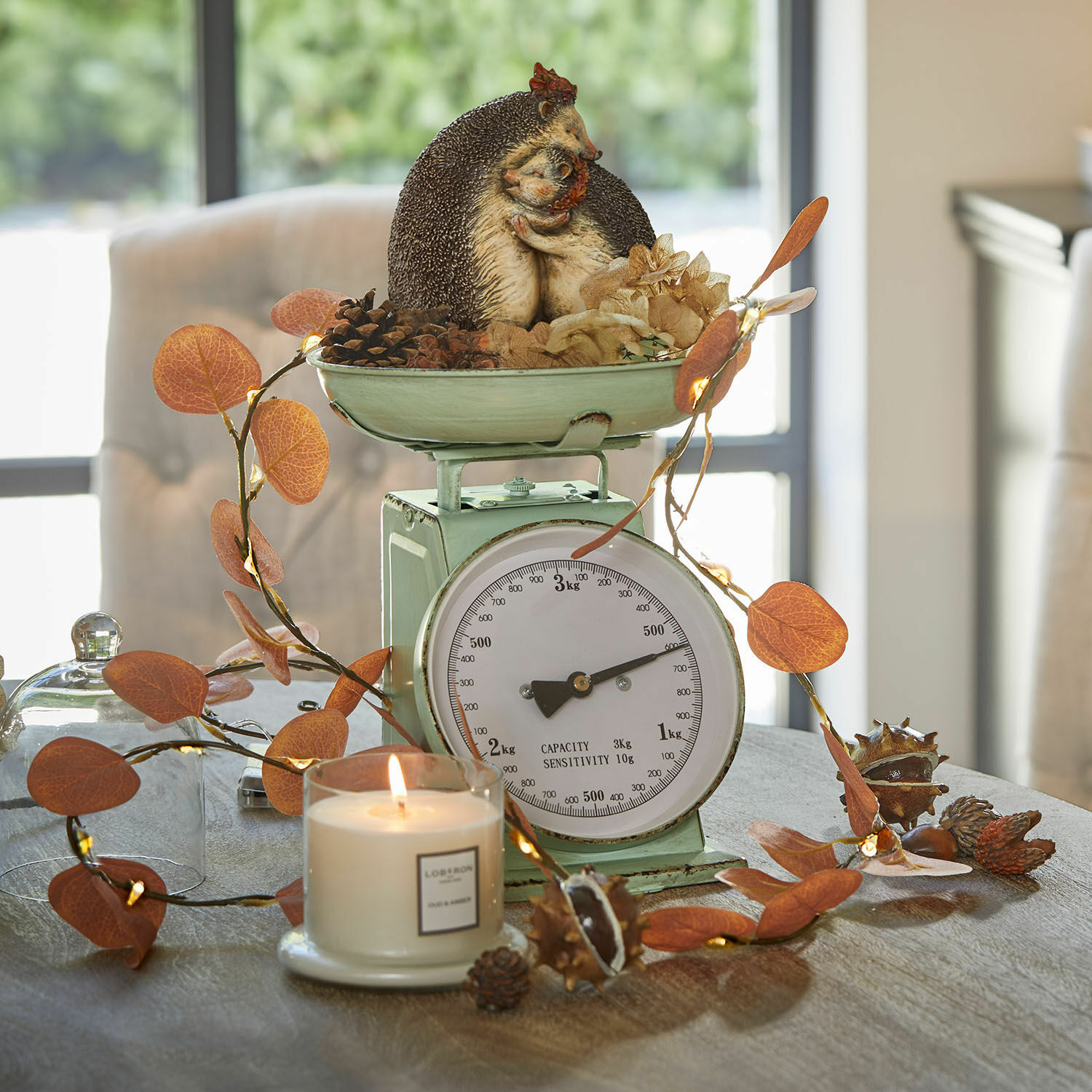 Groene metalen keukenweegschaal met egeldecoratie en oranje bladeren op tafel met brandende kaars