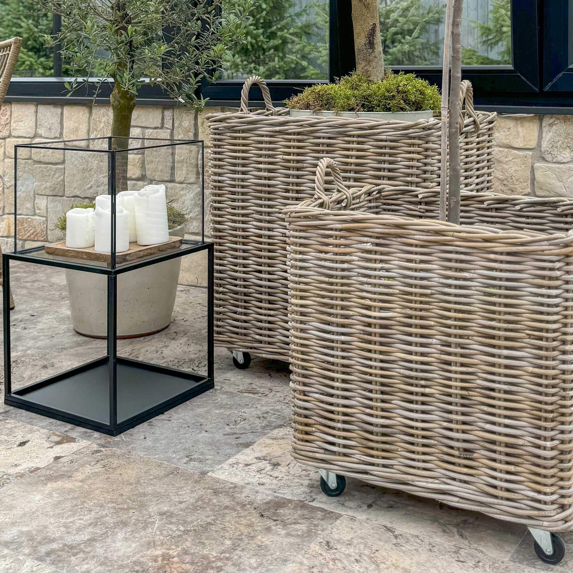 Grote natuurlijke rieten manden op wieltjes op terras naast zwarte tafel met witte kaarsen