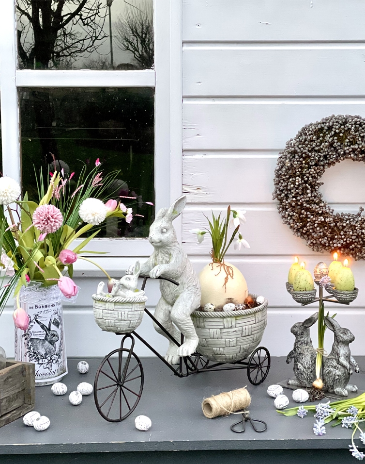 Paasdecoratie met grijze konijnen van metaal en keramiek op tafel met bloemen en kaarsen