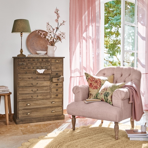 Gezellige roze fauteuil met bloemenkussens naast houten ladekast in lichte woonkamer met roze gordijnen