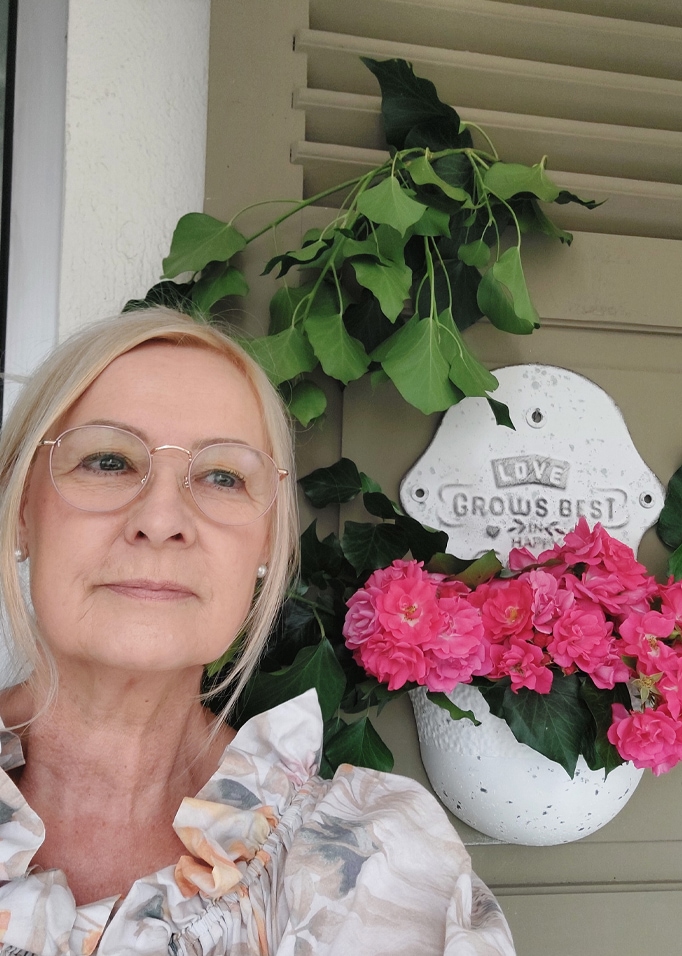 Vrouw met bril naast witte bloempot met roze bloemen en groene bladeren tegen muur