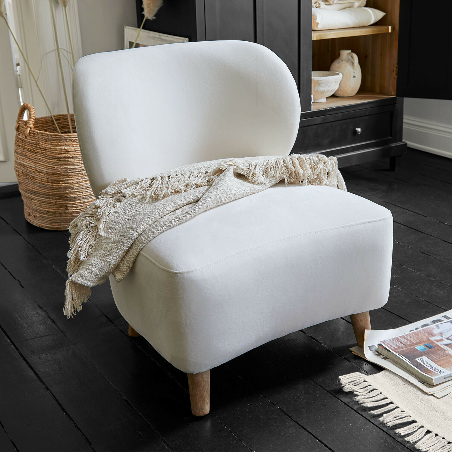 Witte gestoffeerde fauteuil met houten poten en beige deken op zwarte vloer in woonkamer