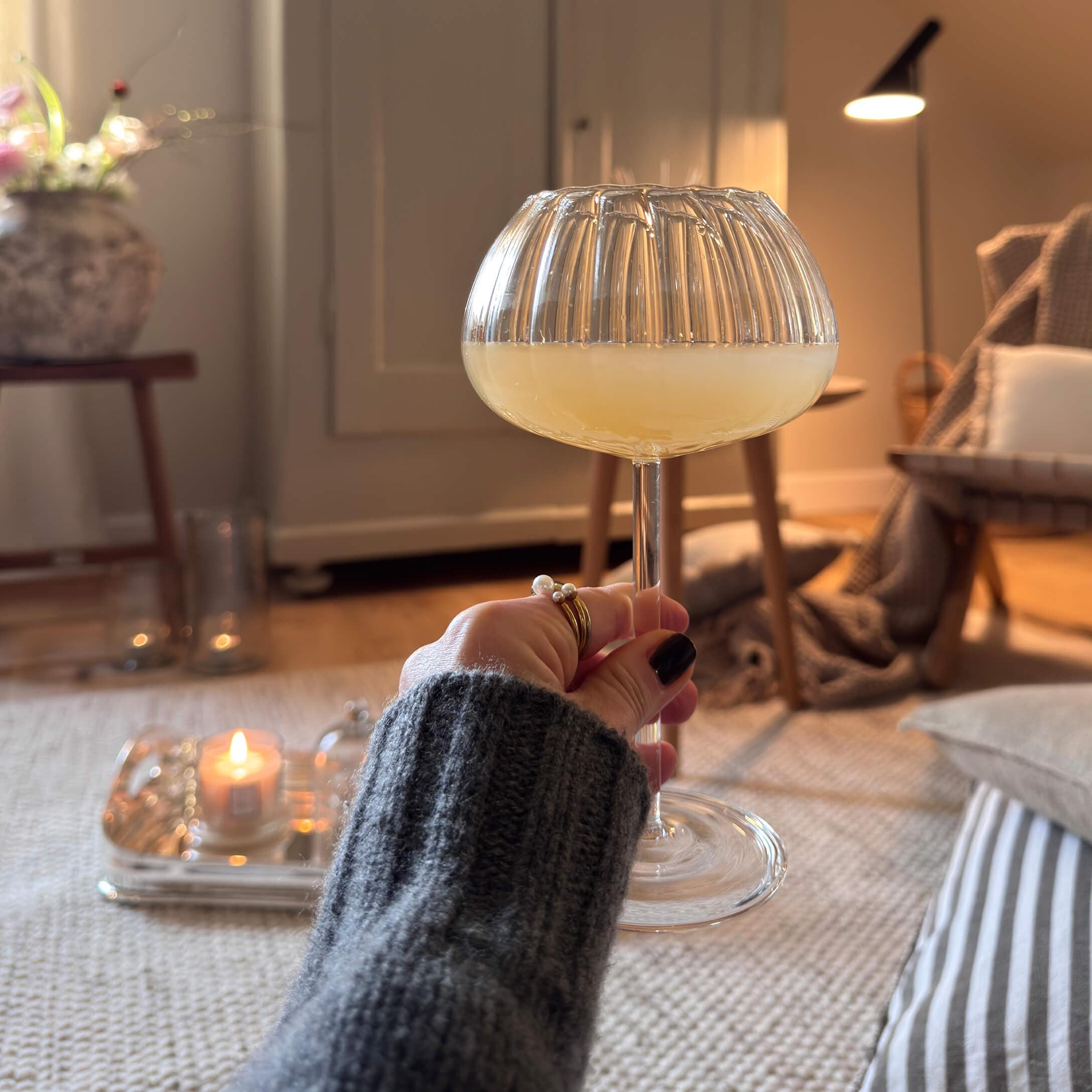 Hand die een glas met gele drank vasthoudt in een gezellige ruimte met zachte verlichting en decoraties
