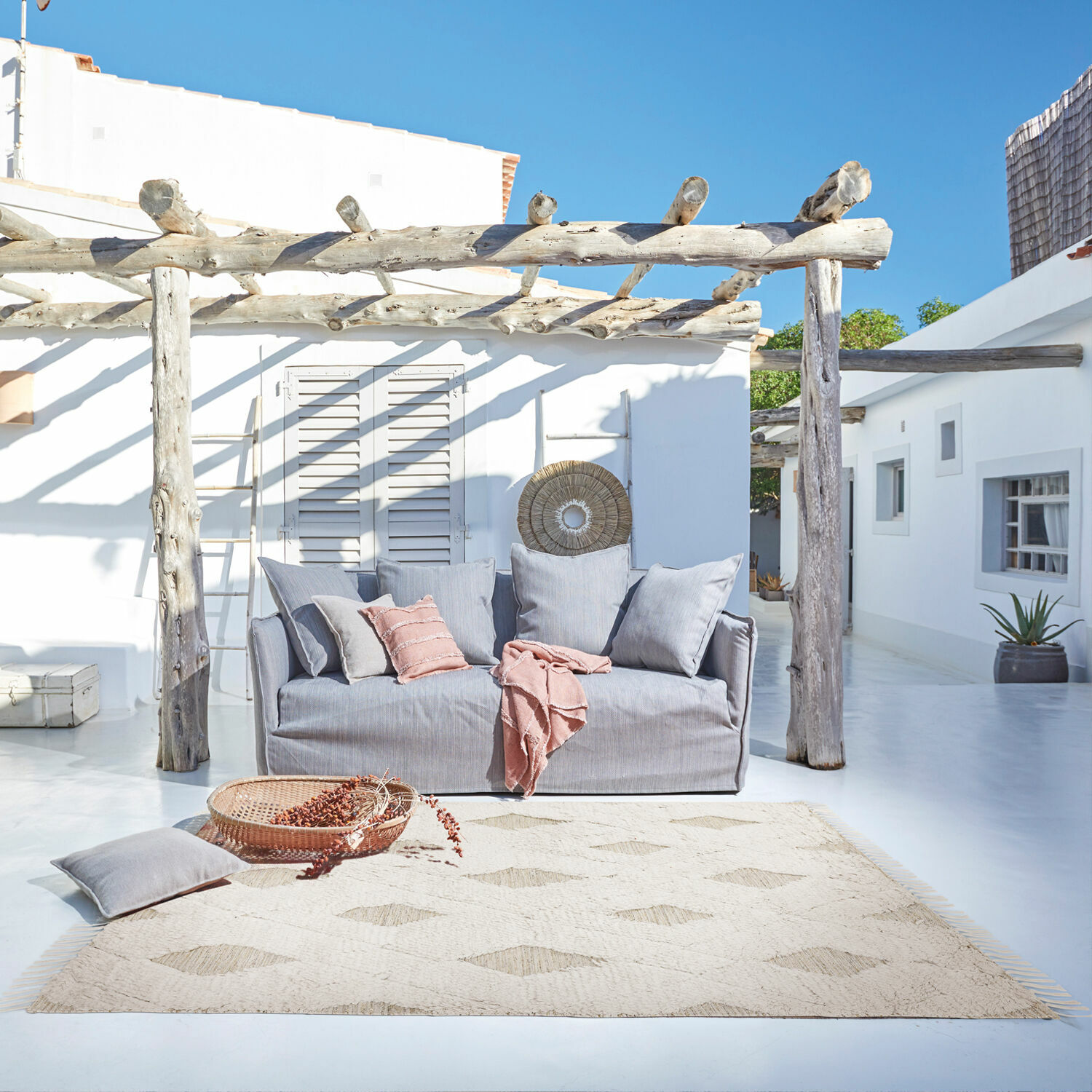 In- & outdoor vloerkleed Meagan Grijze stoffen bank op terras met natuurlijke vloerkleed en roze kussens voor buitenontspanning