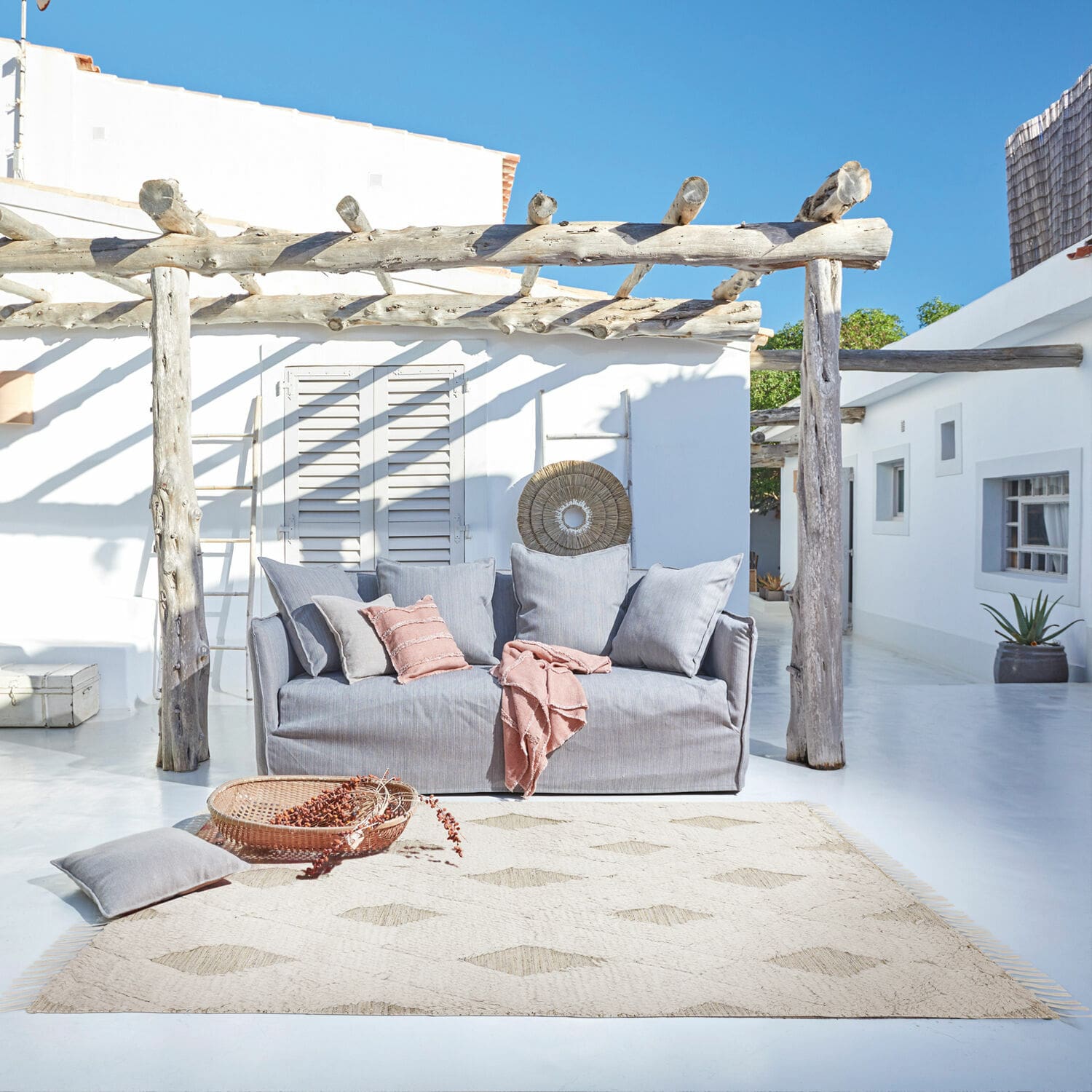 In- & outdoor vloerkleed Meagan Grijze stoffen bank op terras met natuurlijke vloerkleed en roze kussens voor buitenontspanning