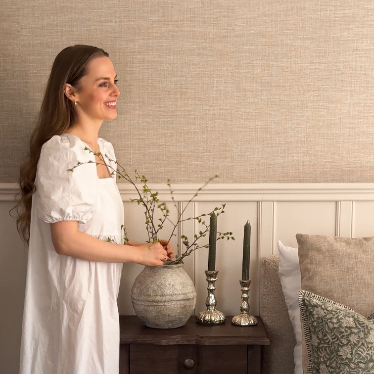 Vrouw in witte jurk rangschikt takken in grijze keramische vaas op houten tafel met kaarsen