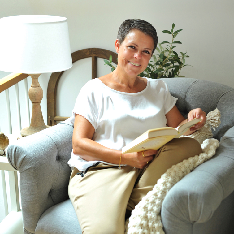 Vrouw in wit shirt en beige broek leest boek op grijze comfortabele bank in lichte kamer
