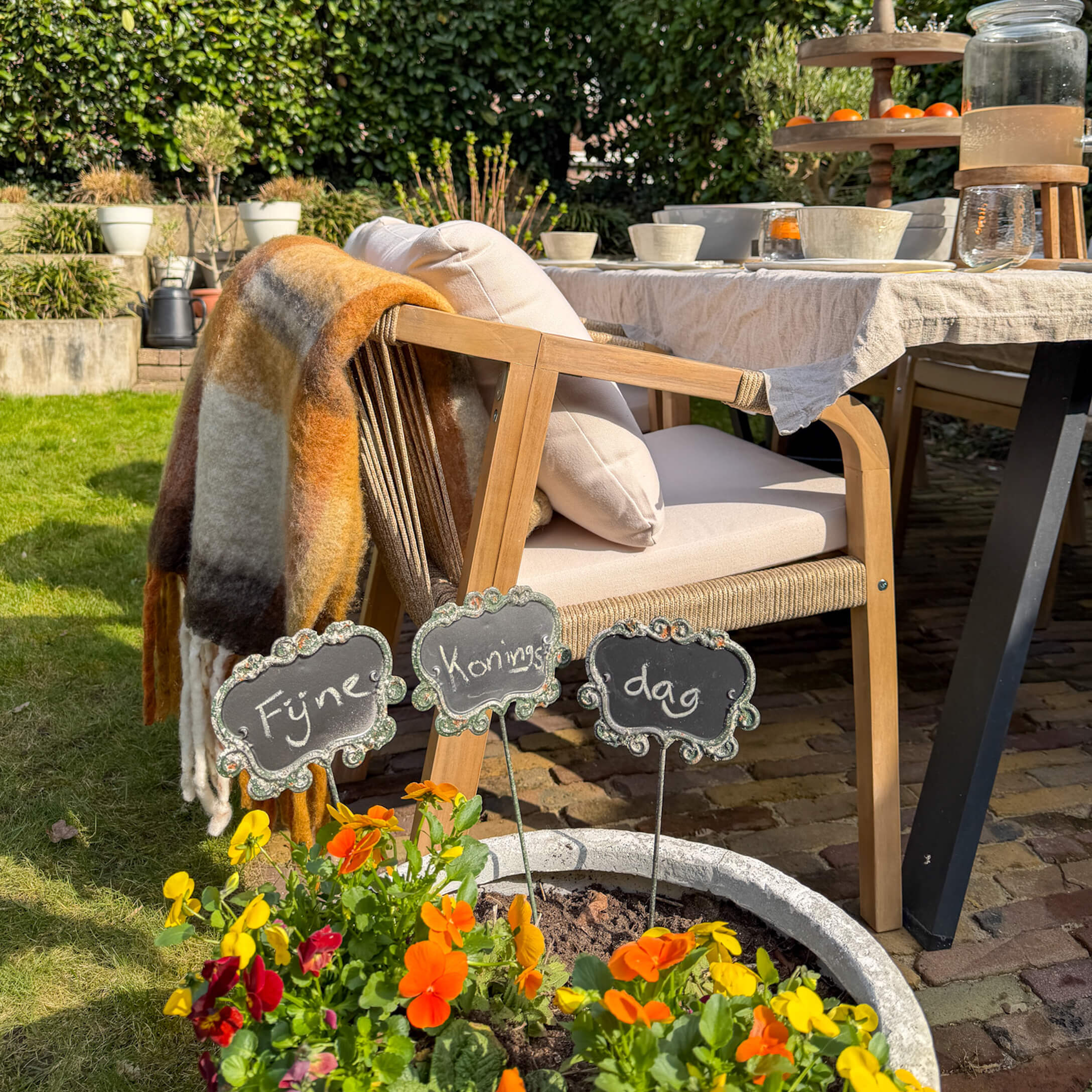Houten tuinstoel met lichte kussens en kleurrijk plaid naast gedekte tafel en bloeiende bloemen in pot
