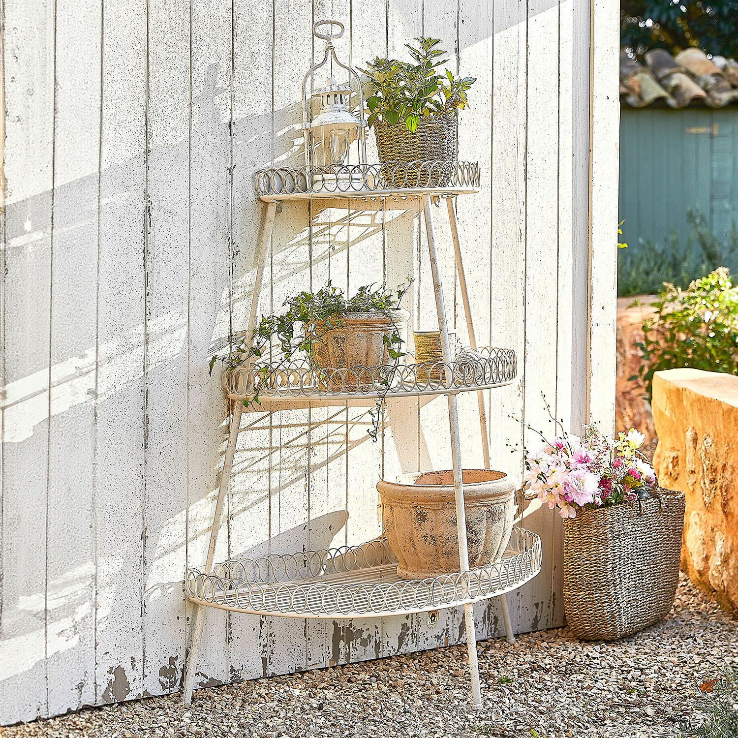 Drie niveaus crème metalen tuinrek met bloempotten en planten tegen een witte muur