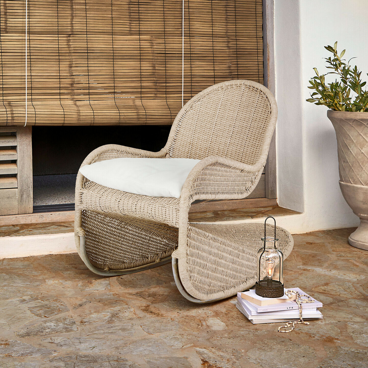 Schommelstoel Fernleigha Beige rotan stoel met wit kussen op stenen vloer bij bamboe rolgordijn