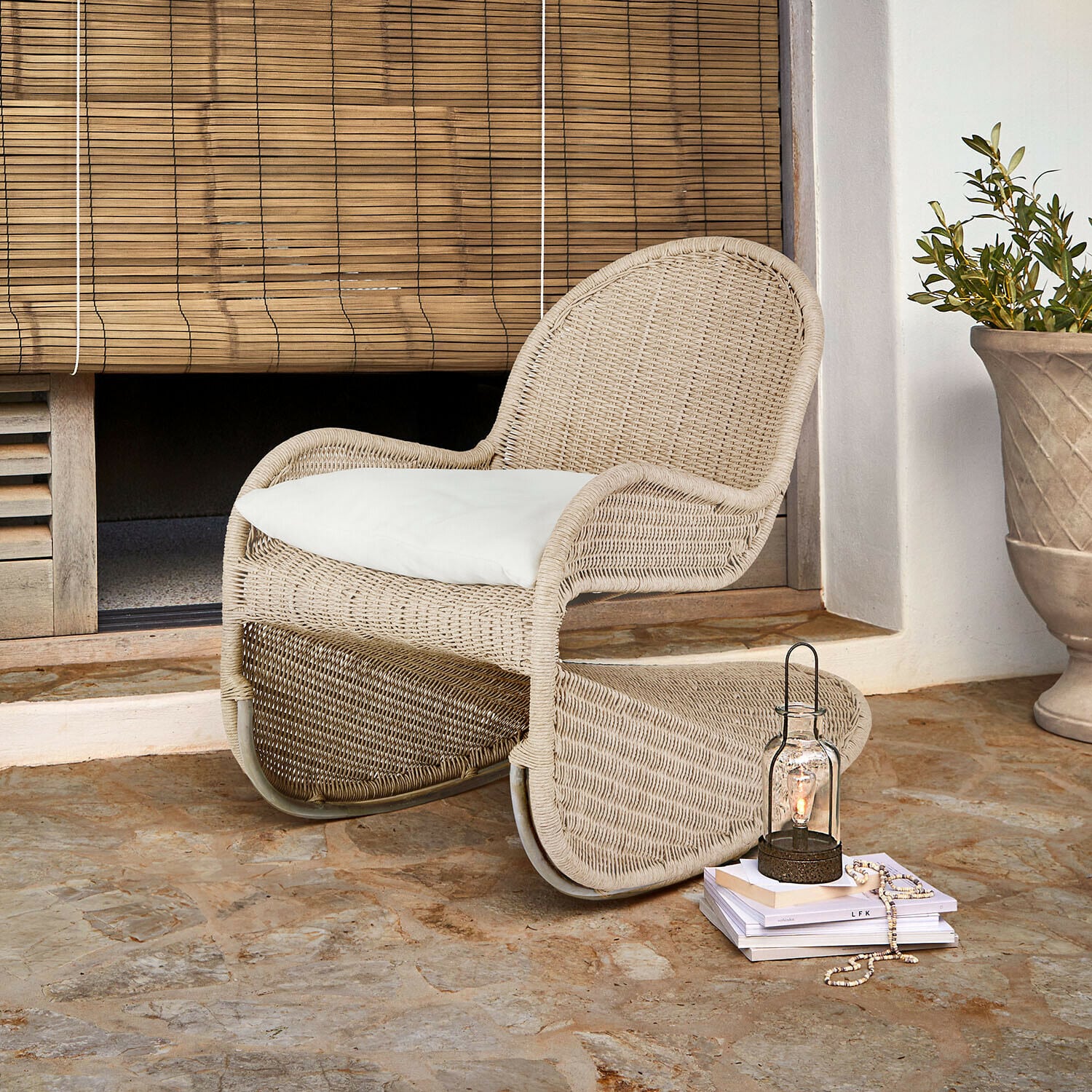 Schommelstoel Fernleigha Beige rotan stoel met wit kussen op stenen vloer bij bamboe rolgordijn