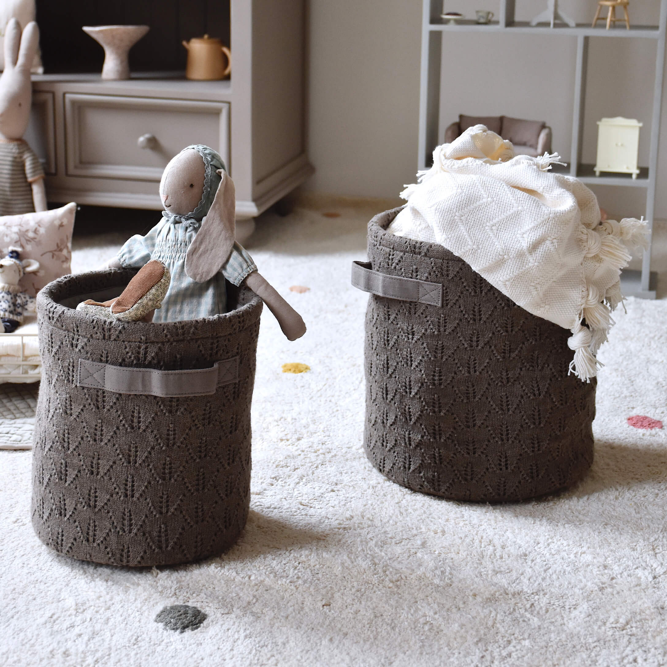 Bruine gebreide opbergmanden met handvatten, gevuld met speelgoed en deken, op een lichte vloerbedekking