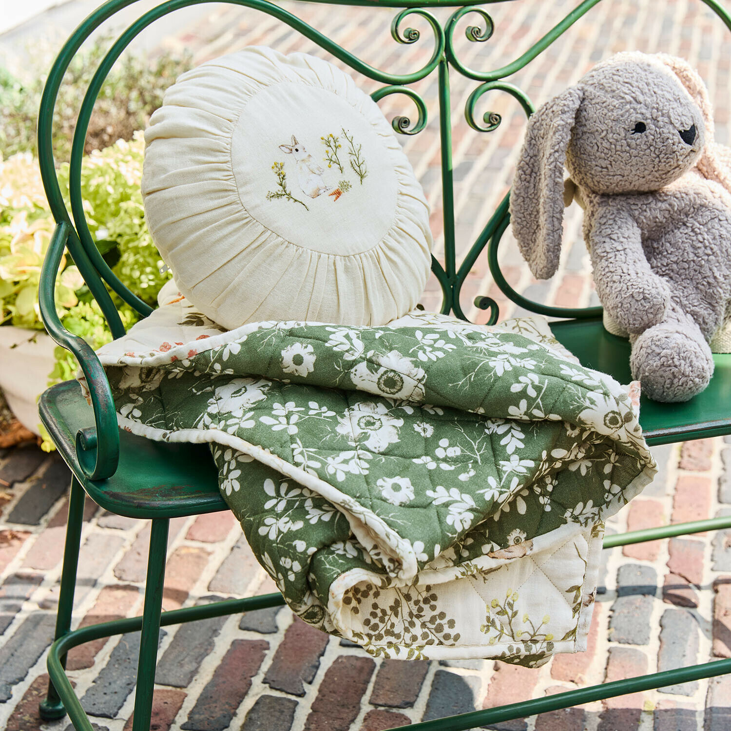 Groen doorgestikt deken met wit bloemenpatroon op groene metalen bank met ronde kussen en knuffelkonijn