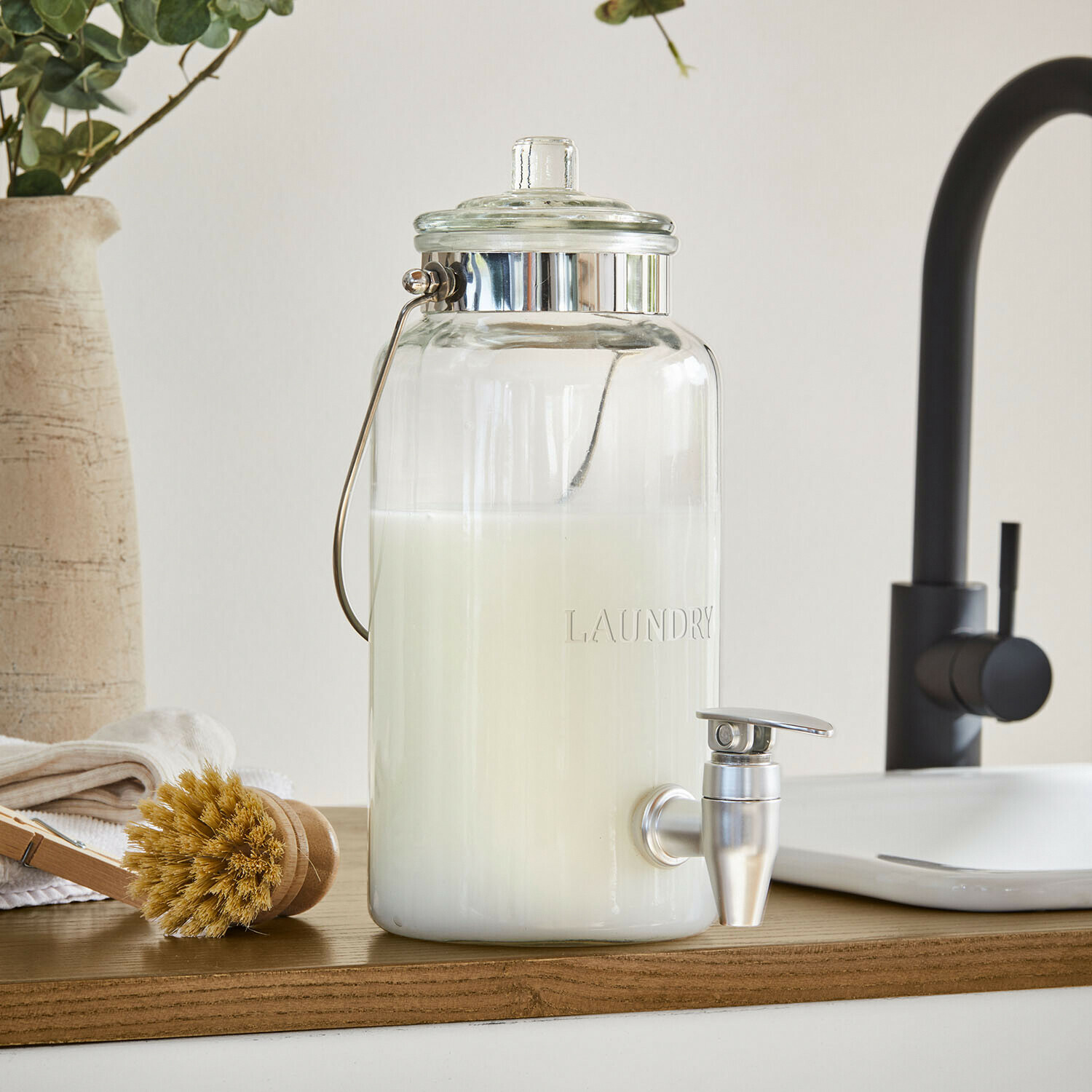 Glazen dispenser voor wasmiddel met metalen kraan en handvat, witte vloeistof binnenin