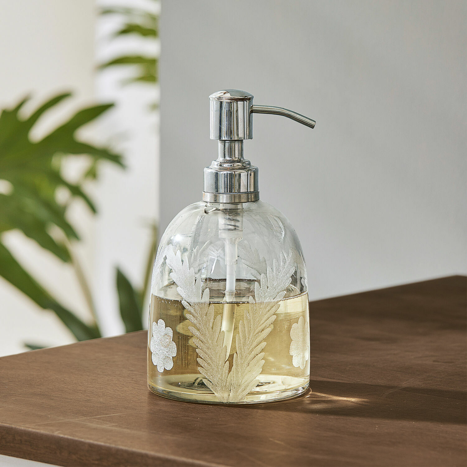 Zeepdispenser Crystal Glazen zeepdispenser met metalen pomp en wit bladmotief op bruine tafel