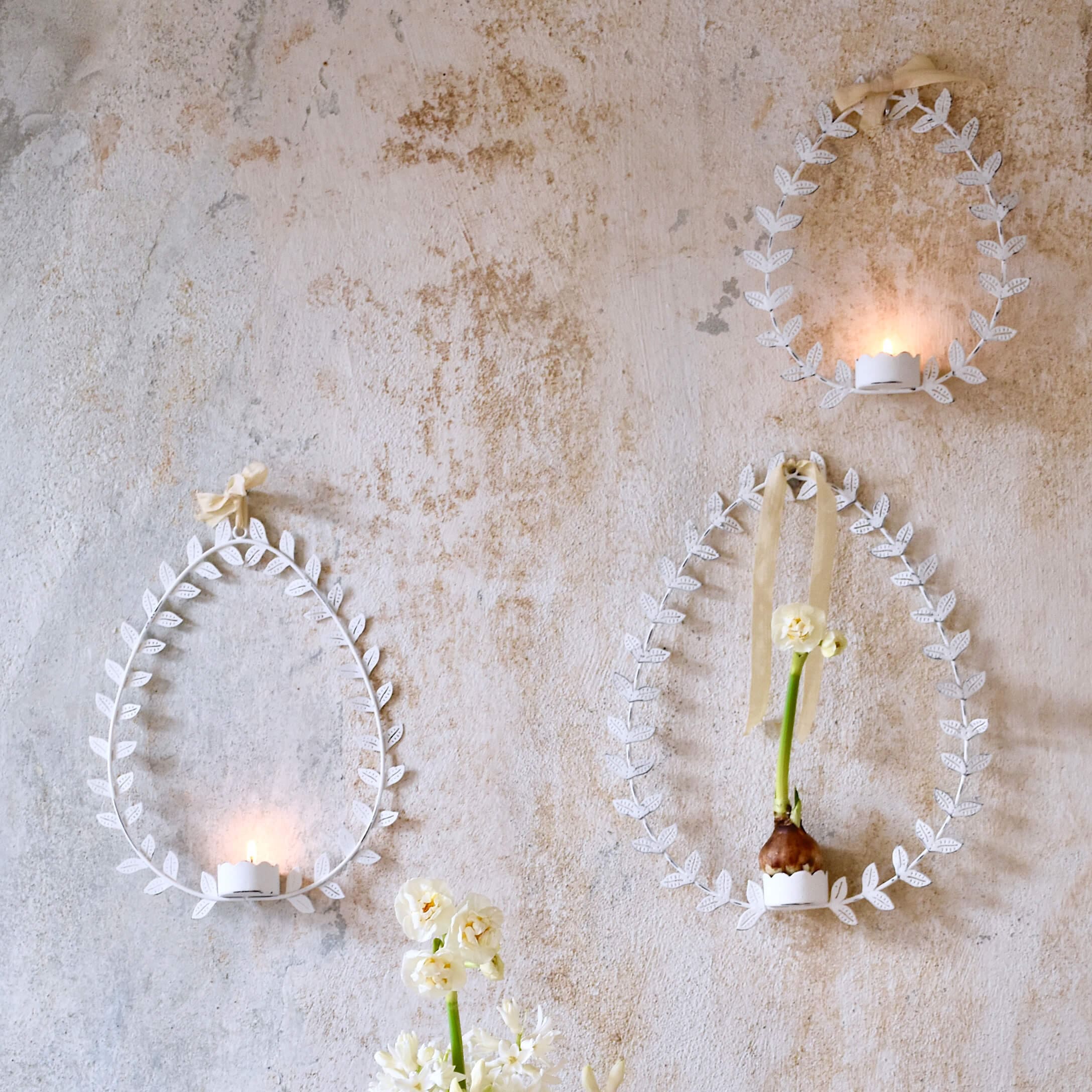 Witte metalen wanddecoraties in eivorm met bladeren, kandelaren en bloemen op pastelkleurige muur