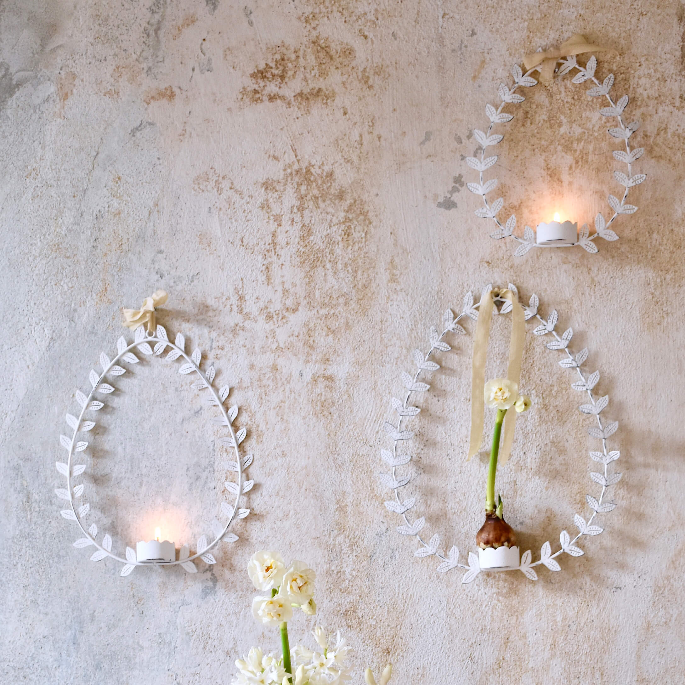 Witte metalen wanddecoraties in eivorm met bladeren, kandelaren en bloemen op pastelkleurige muur