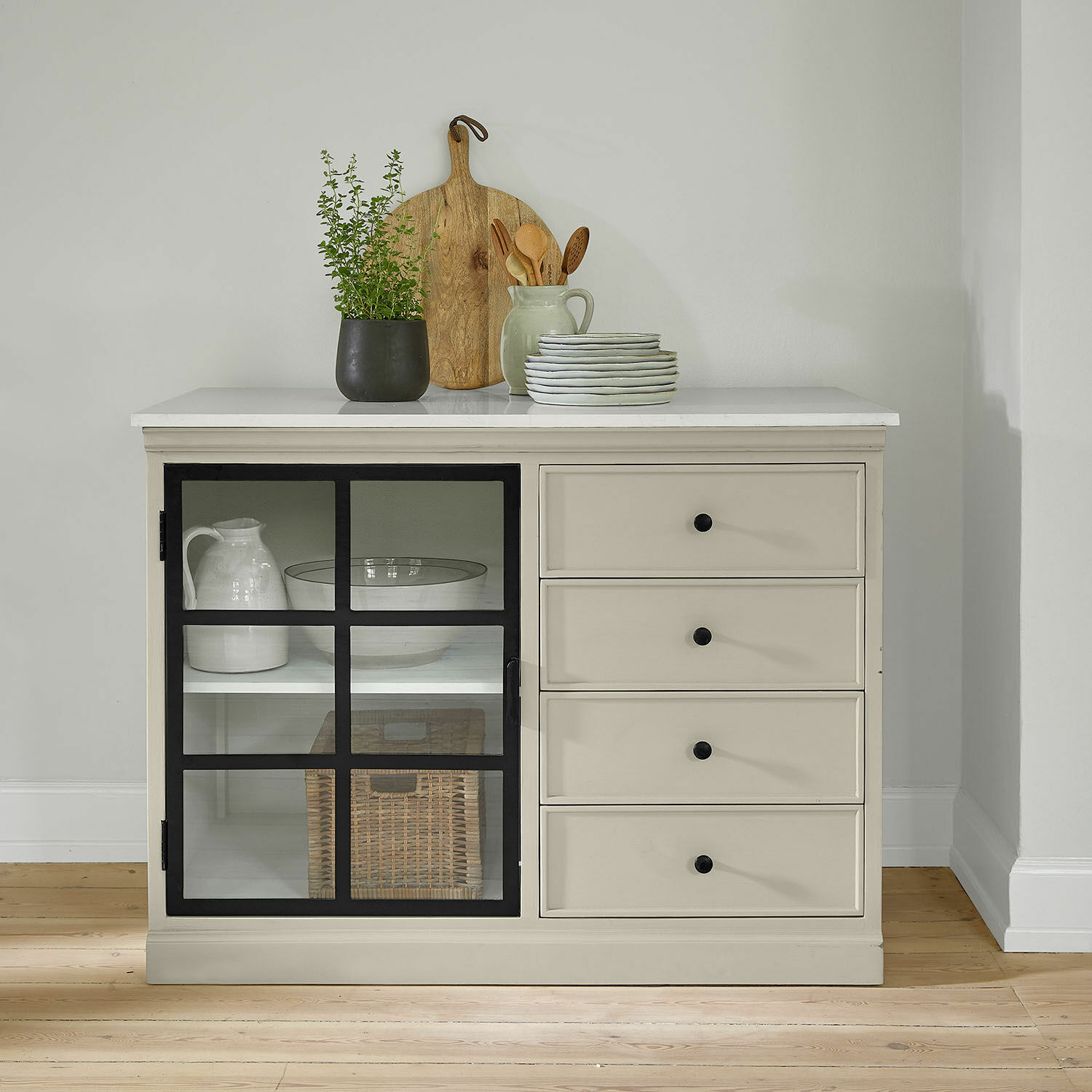 Beige dressoir met zwarte handgrepen en glazen deur, gedecoreerd met houten plank en groene plant