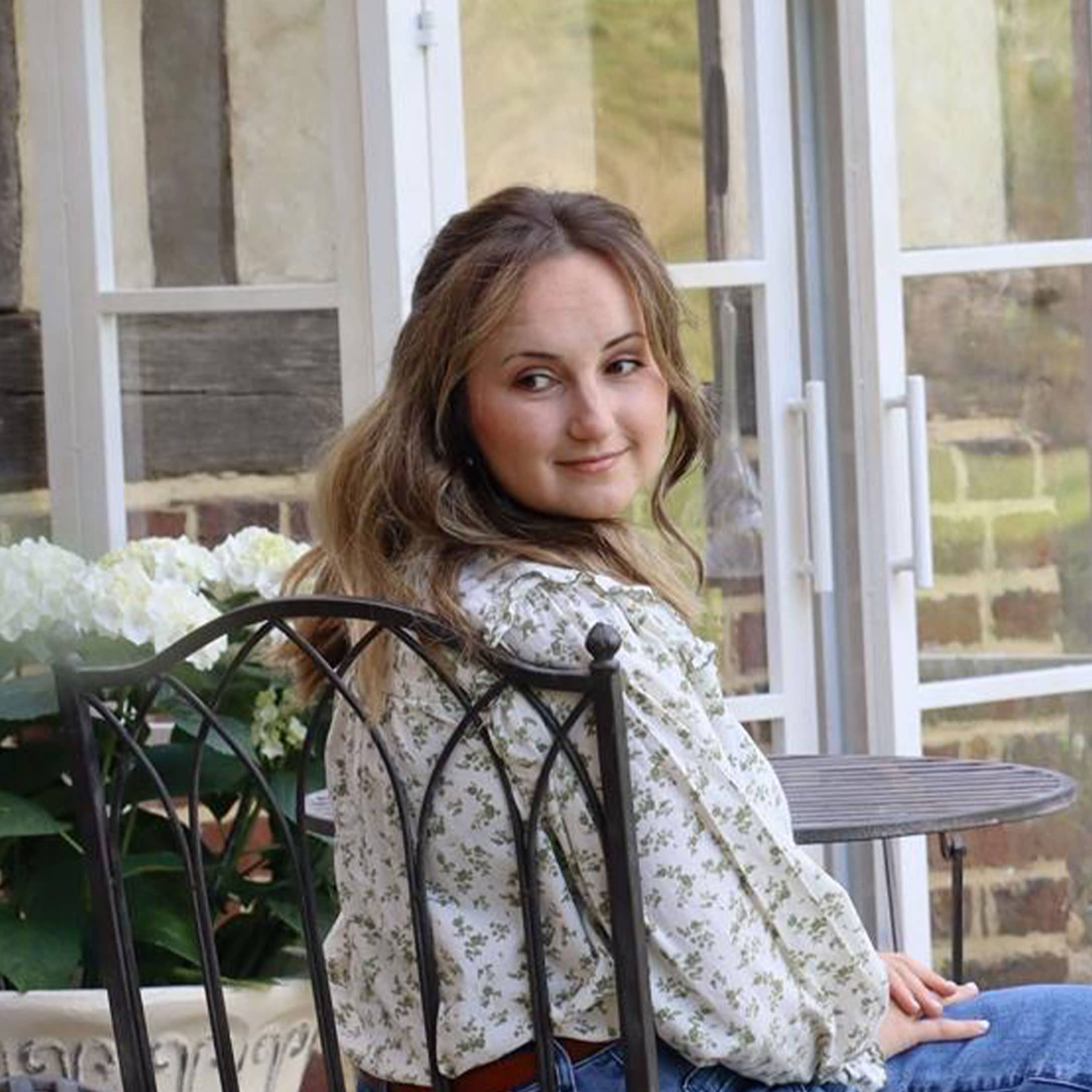 Vrouw in gebloemde blouse zittend op metalen stoel in lichte tuin met witte bloemen