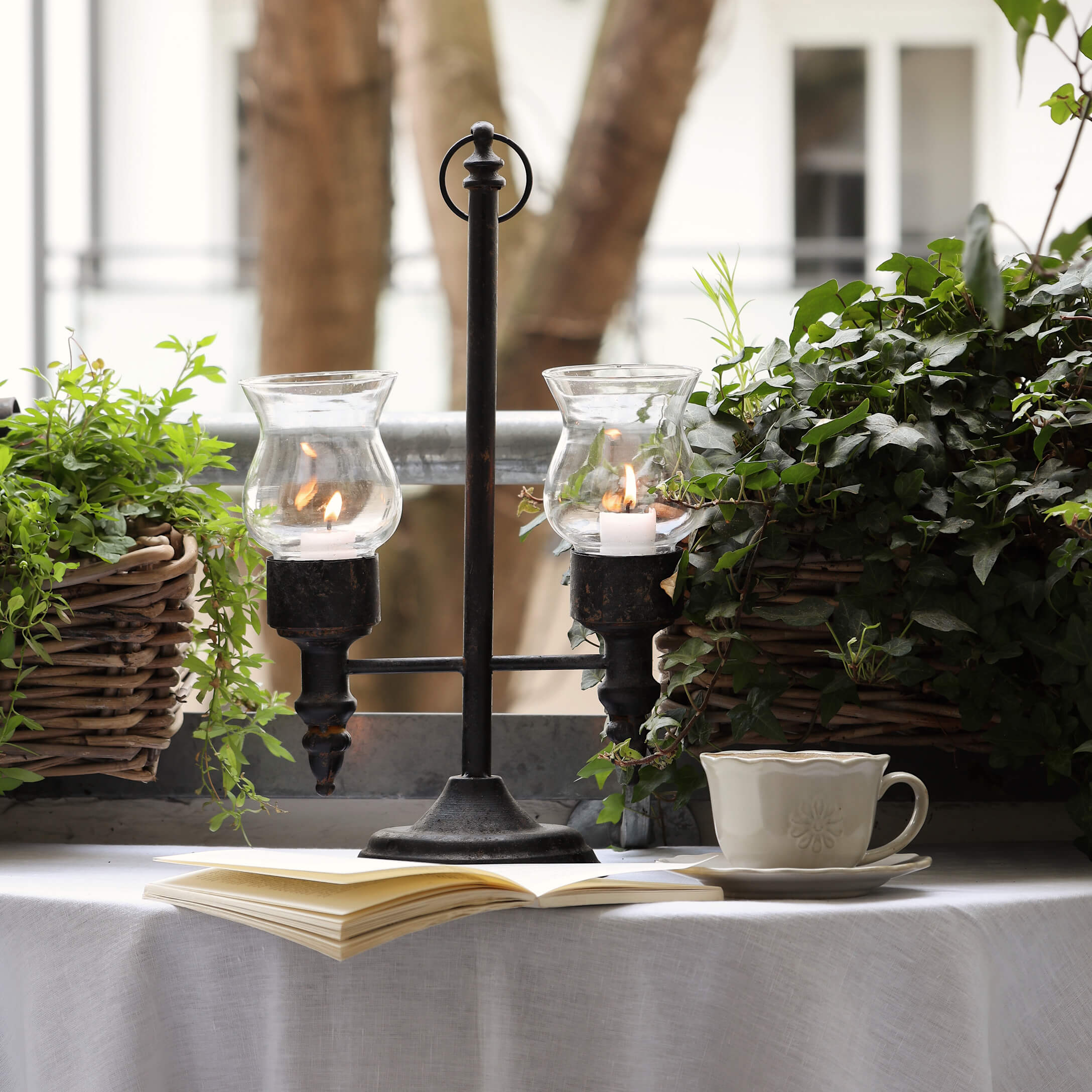 Metalen kandelaar met twee glazen kappen tussen groene planten op balkon