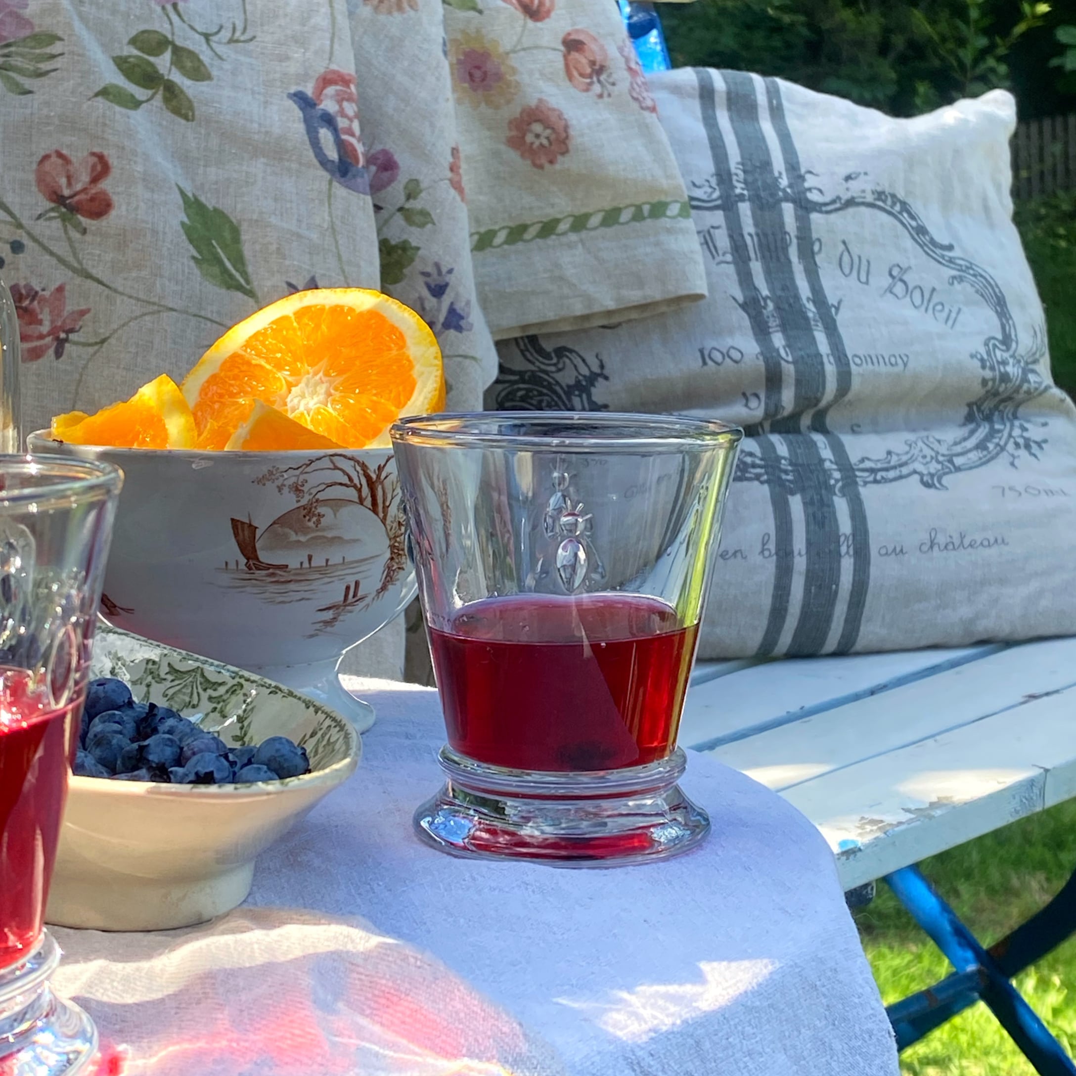 Glas met rode drank op wit tafelkleed, naast kom met sinaasappels en bosbessen buiten