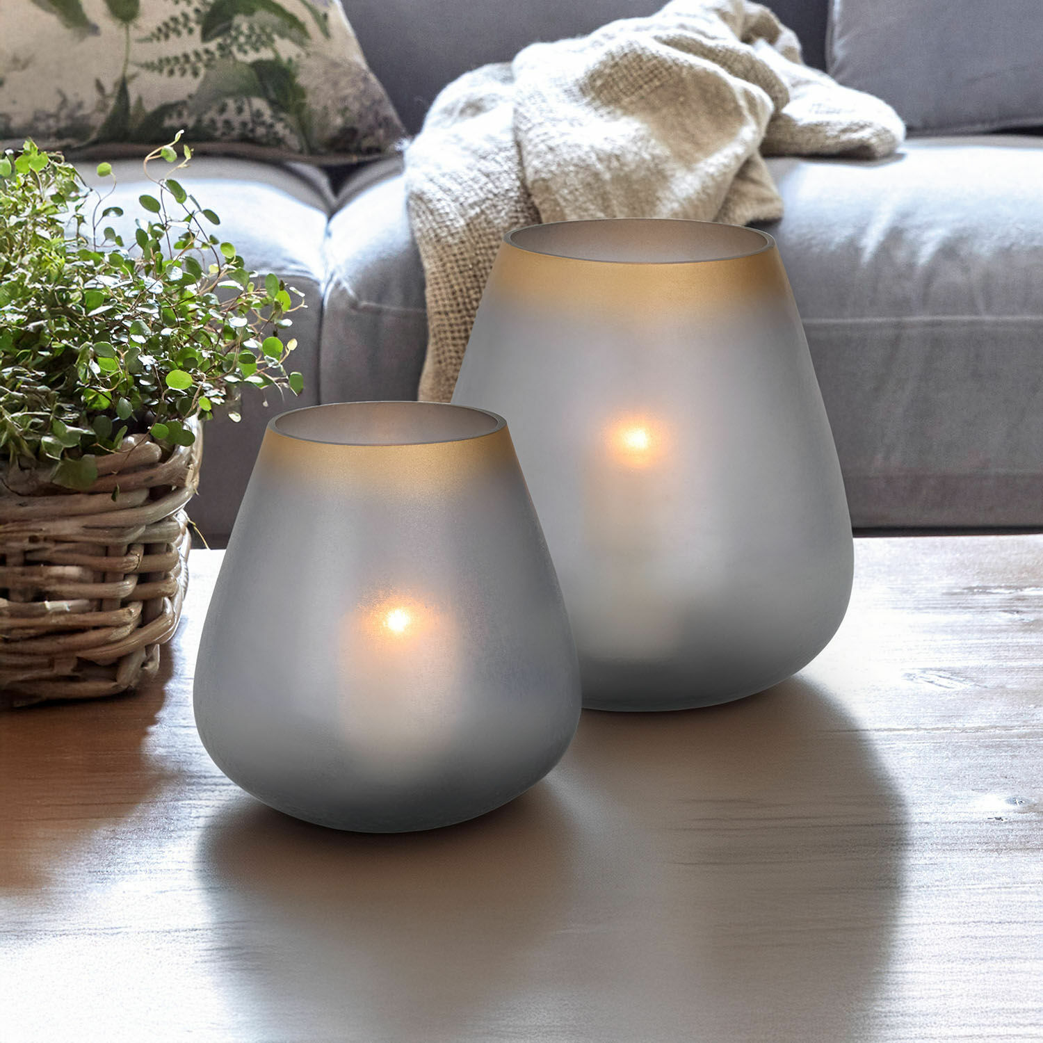 Twee matgrijze glazen theelichthouders op houten tafel in woonkamer met plant en bank