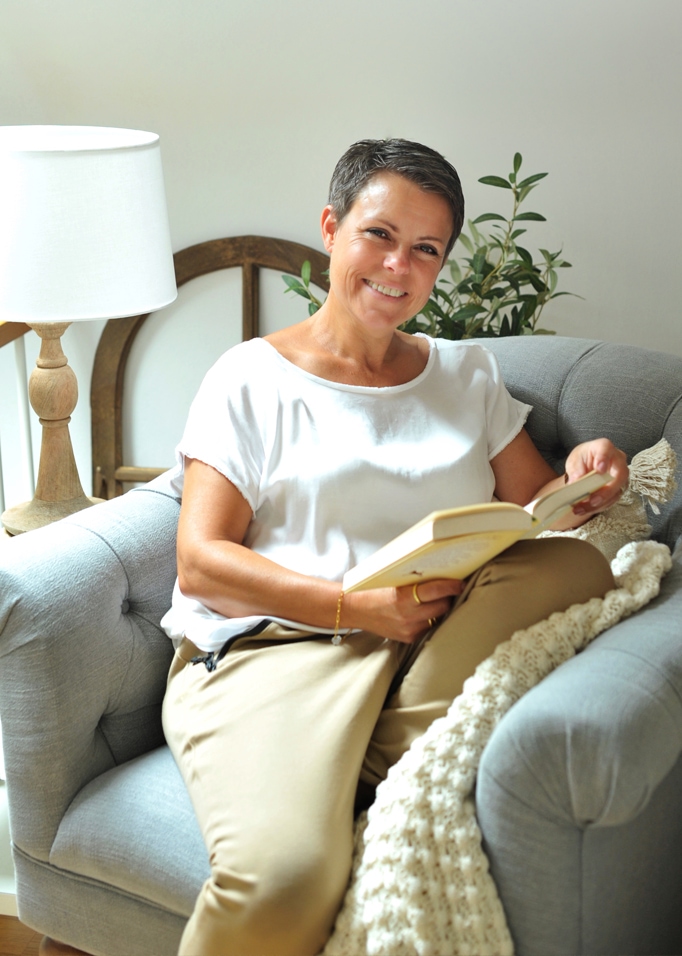 Vrouw leest boek op een grijze gestoffeerde bank met witte deken en houten lamp ernaast