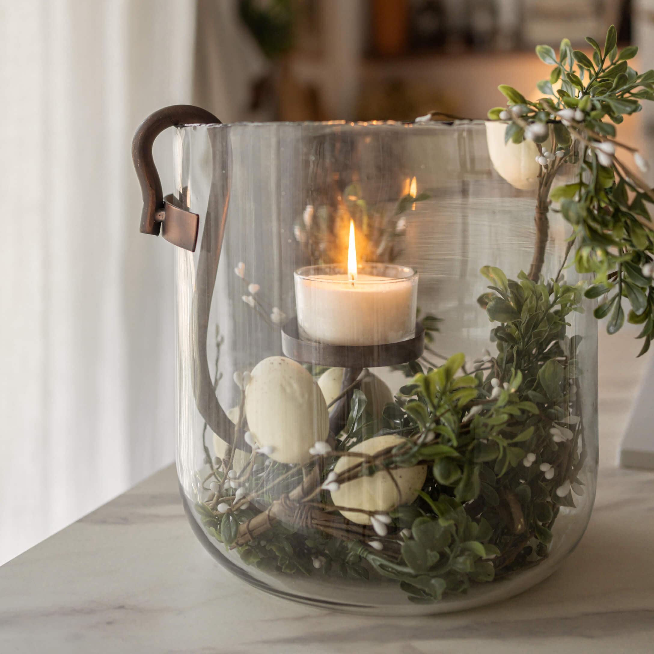 Glazen lantaarn met metalen handvat, brandende witte kaars en groene decoratie met witte bessen