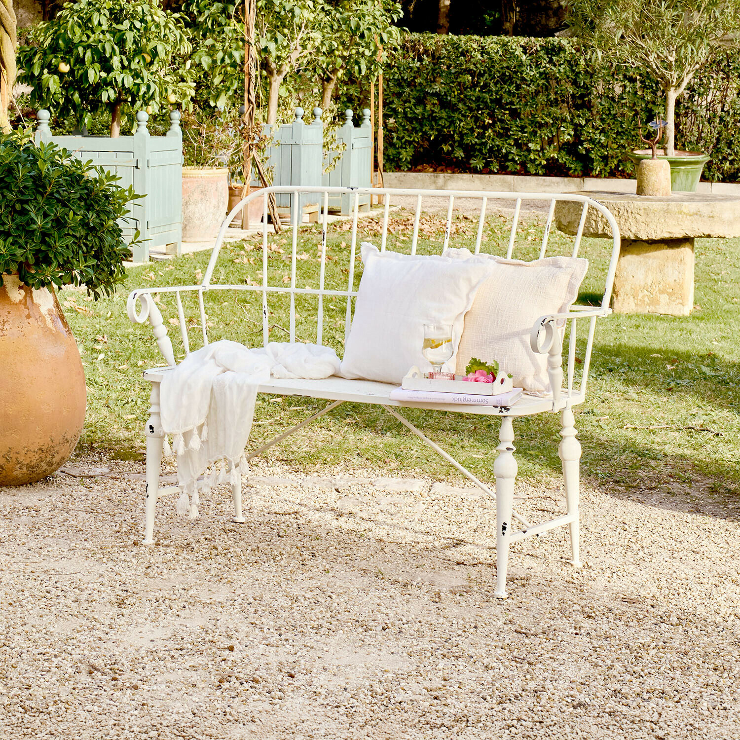 Witte metalen tuinbank met kussens en deken op grindterras in groene tuin