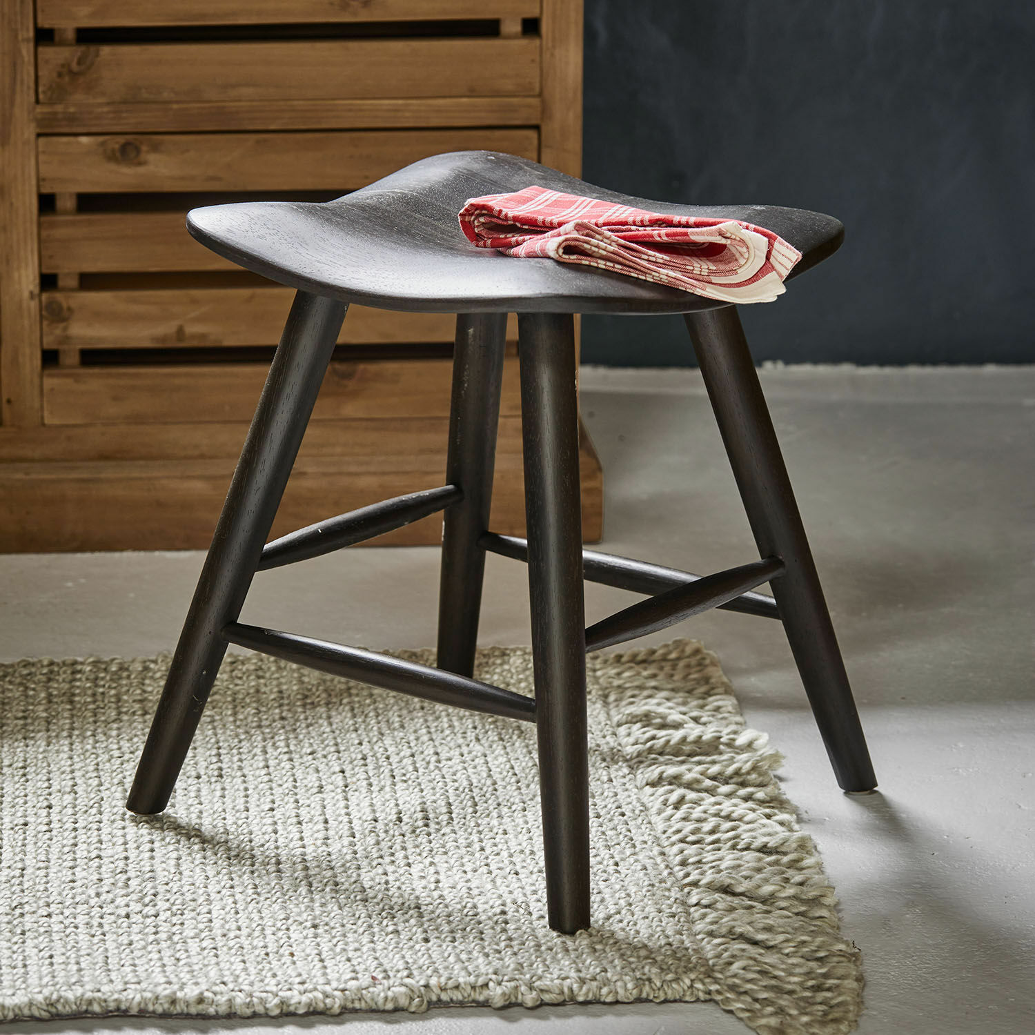 Zwarte houten kruk met gebogen zitting op lichte vloerkleed, gevouwen rood decoratief doek