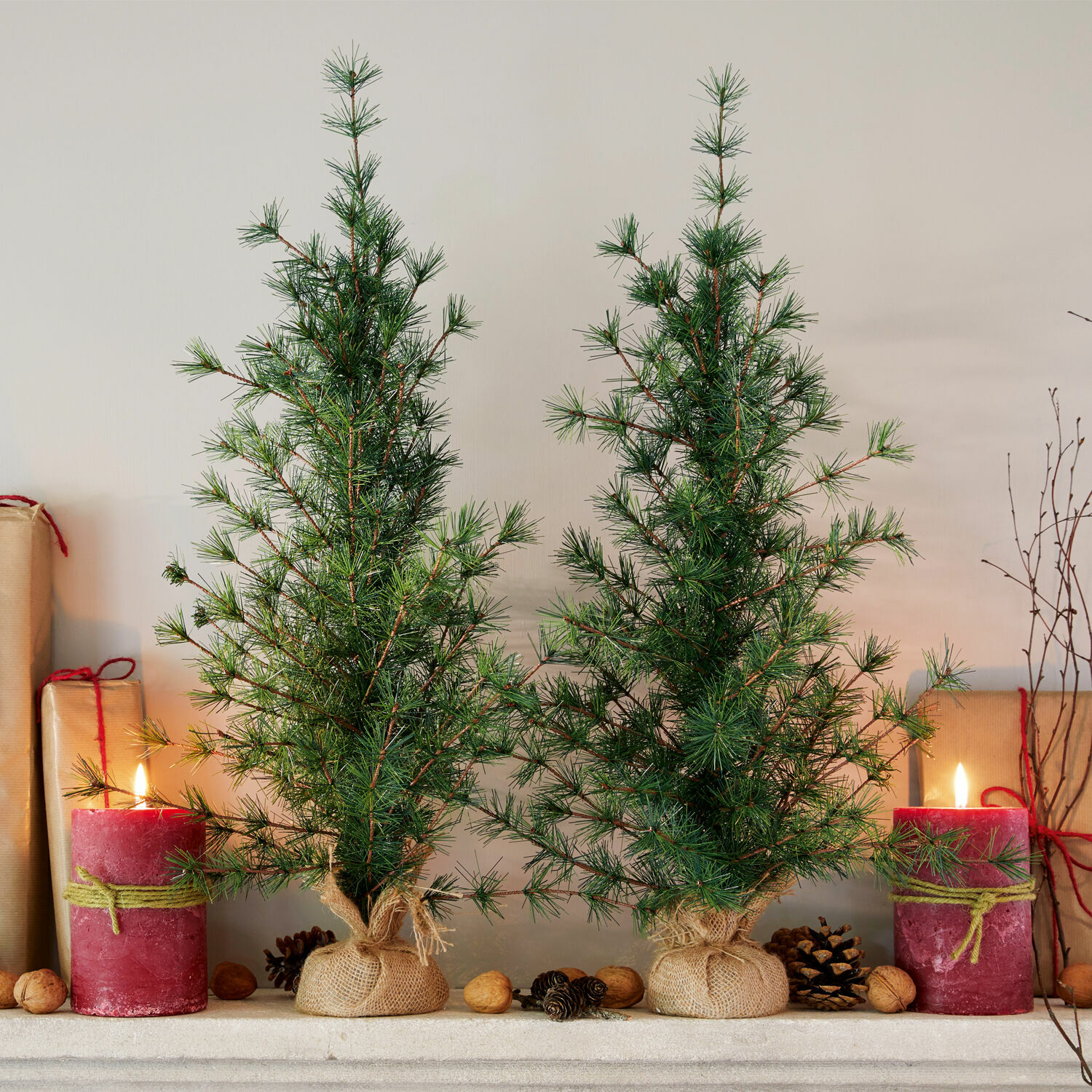 Sierboom set van 2 Bohicon Twee kleine groene kerstbomen in jute zakken met rode kaarsen en noten op de schoorsteenmantel