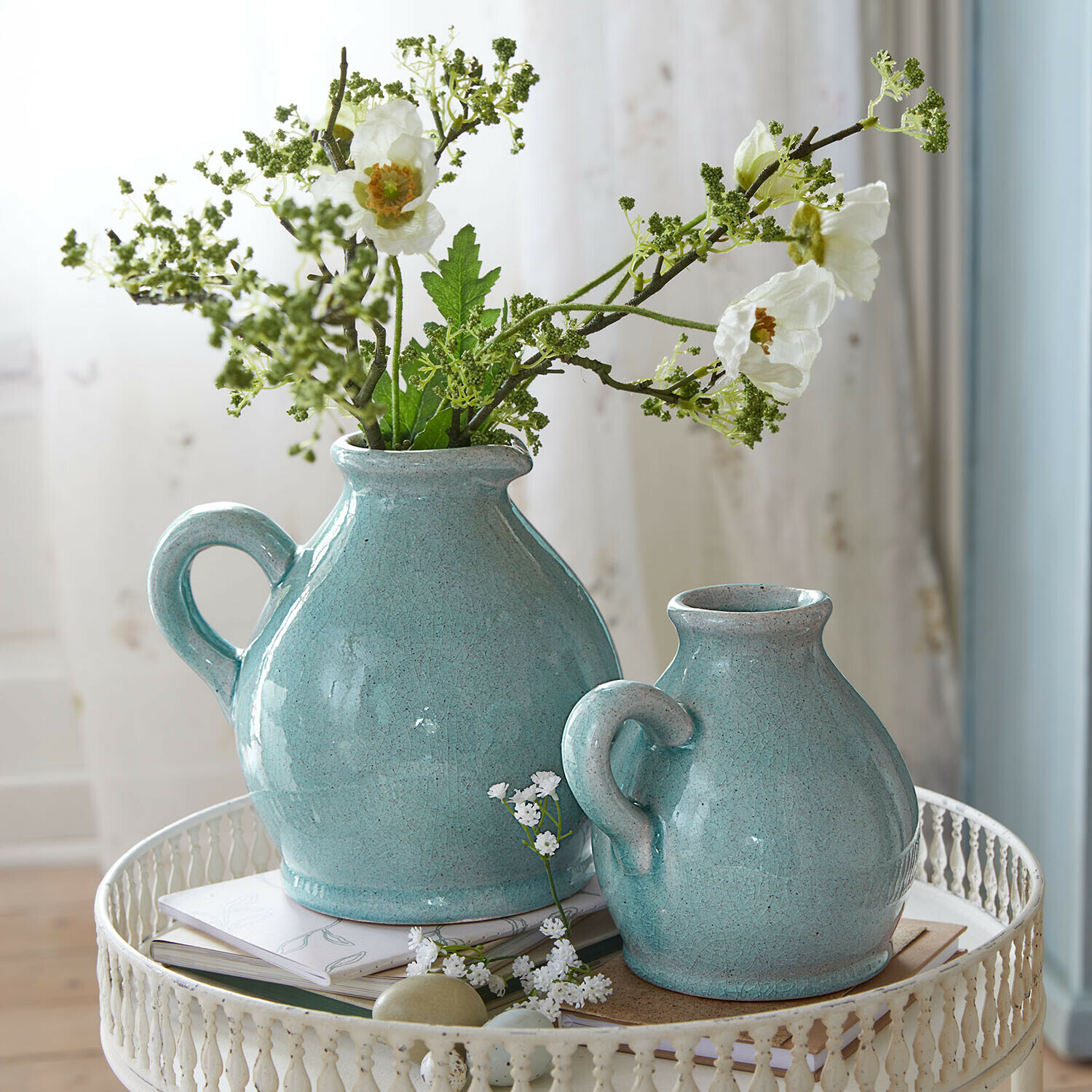 Twee turquoise keramische kannen met handvatten op een witte metalen tafel met bloemen en boeken