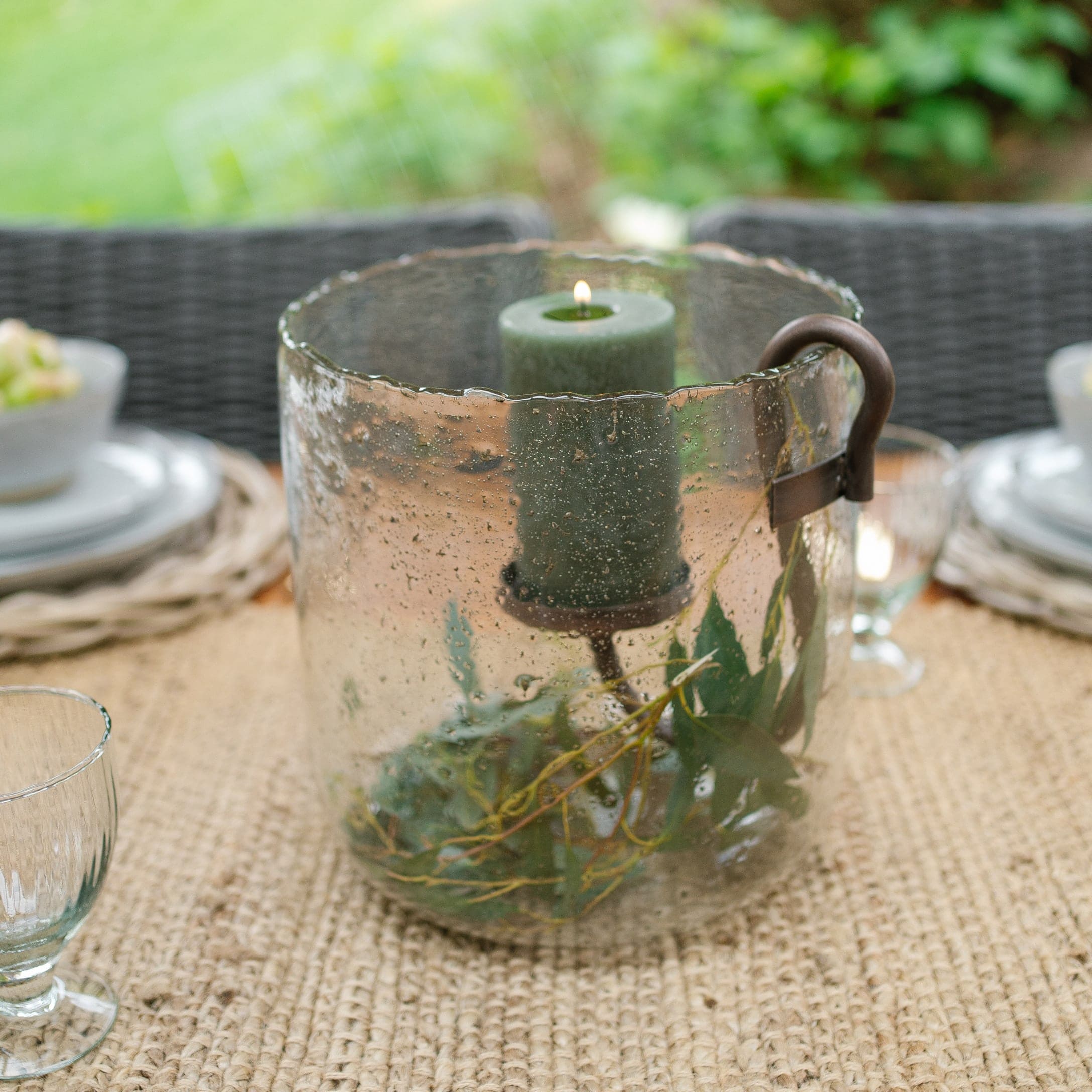 Glazen windlicht met groene kaars en metalen handvat op rustieke tuintafel